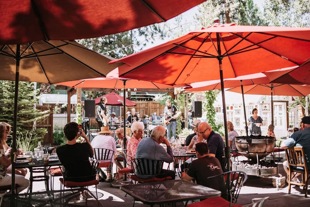 An outdoor courtyard with metal tables set up underneath red umbrellas. Groups of diners are sitting at the tables in the background.