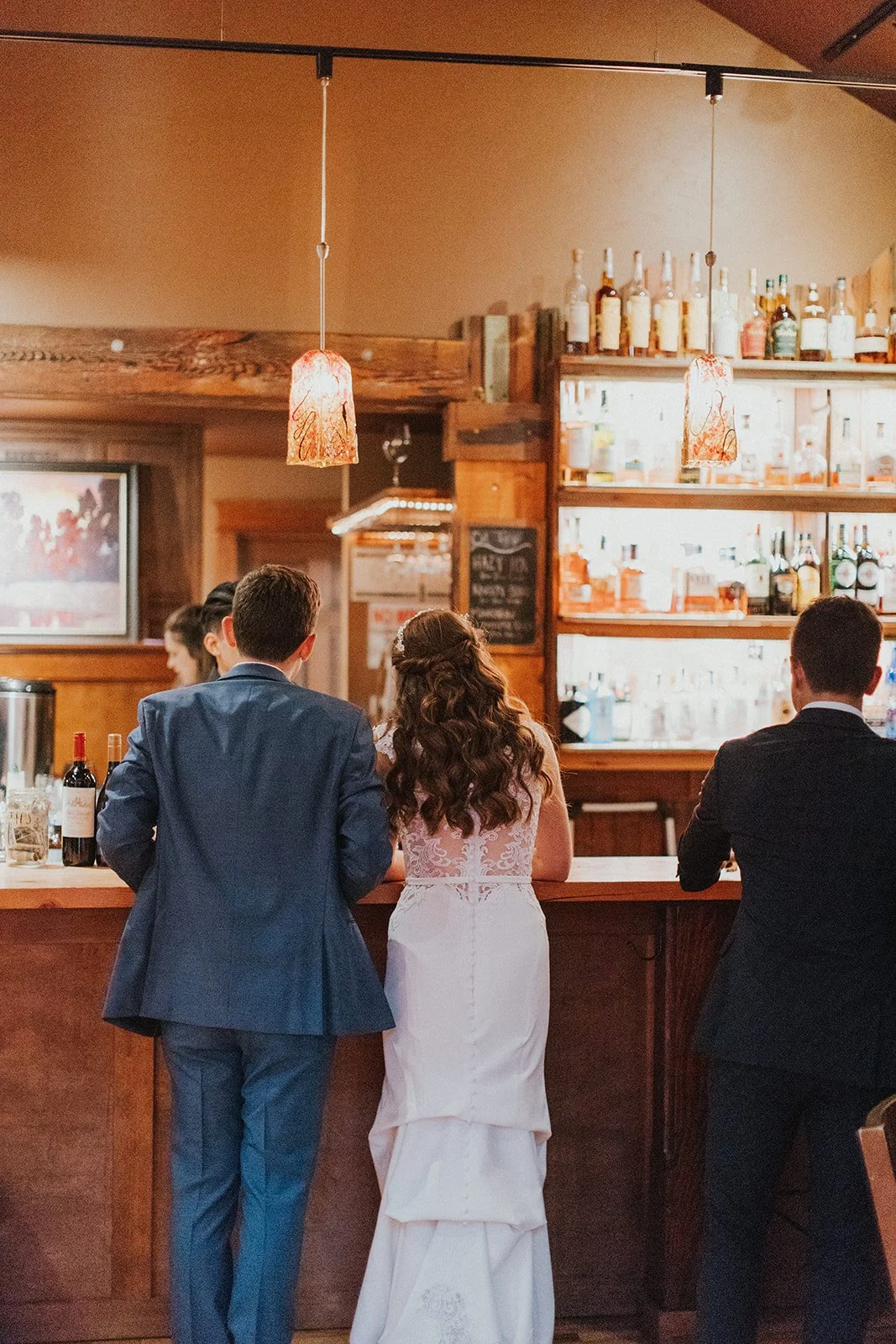 A woman in a white wedding dress and a man in a blue suit stand at a wooden bar, facing away from the camera. In the background are shelves lit up with bright bottles and soft, warm indoor lighting.