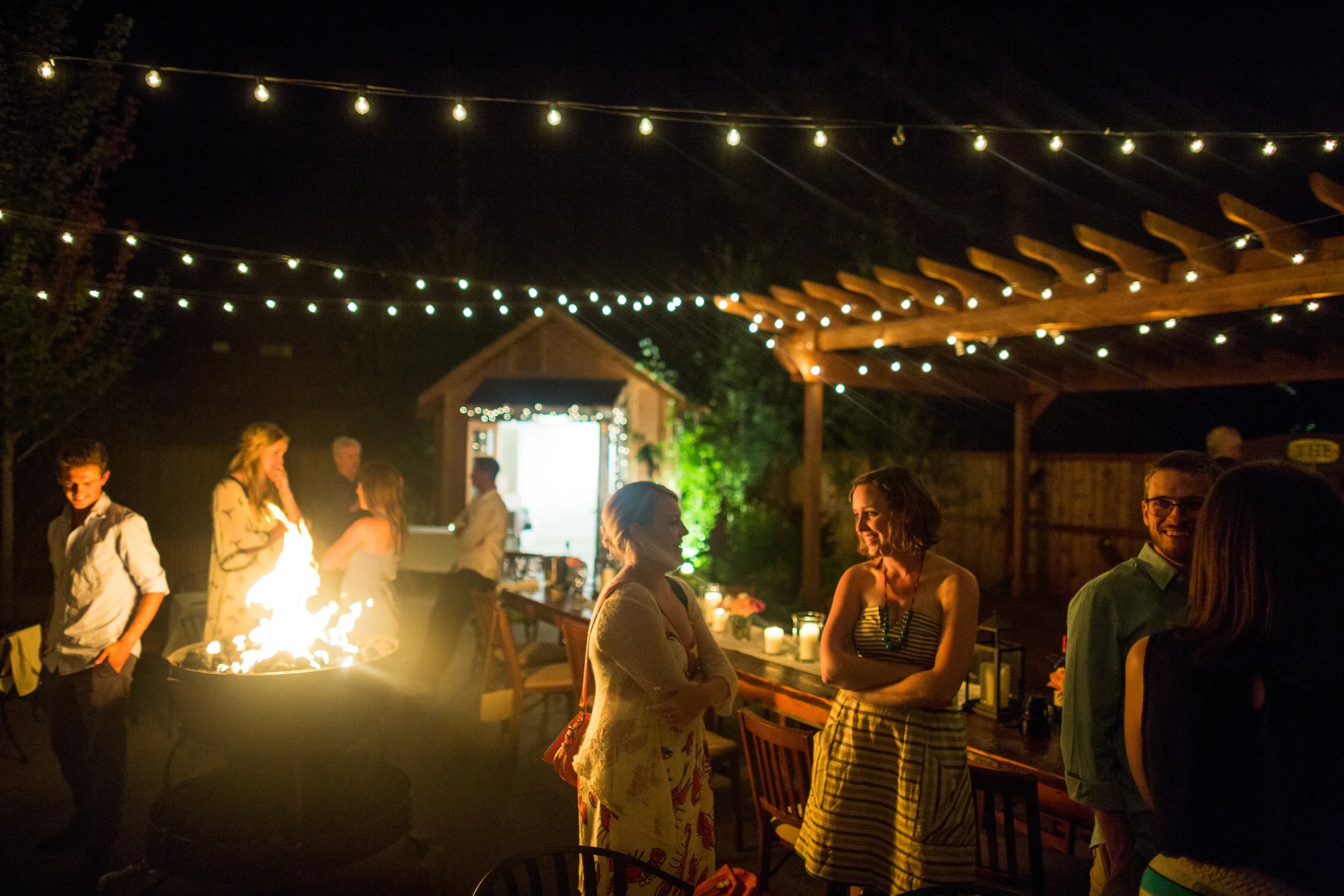Two women chatting and smiling in an outdoor courtyard amongst small groups under string lights. It's nighttime, with a bright fire pit and tables and chairs in the background.