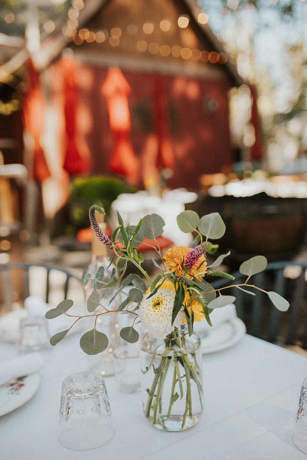 A close-up of flowers and leaves in a clear glass vase set on top of a white table cloth with place settings in an outdoor courtyard. The background is blurry, with a barn red building in the distance.