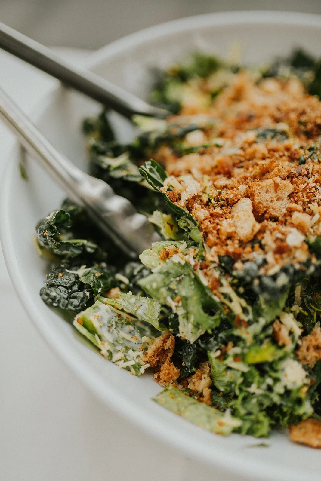 A close-up of a pair of silver tongs in a bowl of Caesar salad. The salad is topped with crushed croutons.