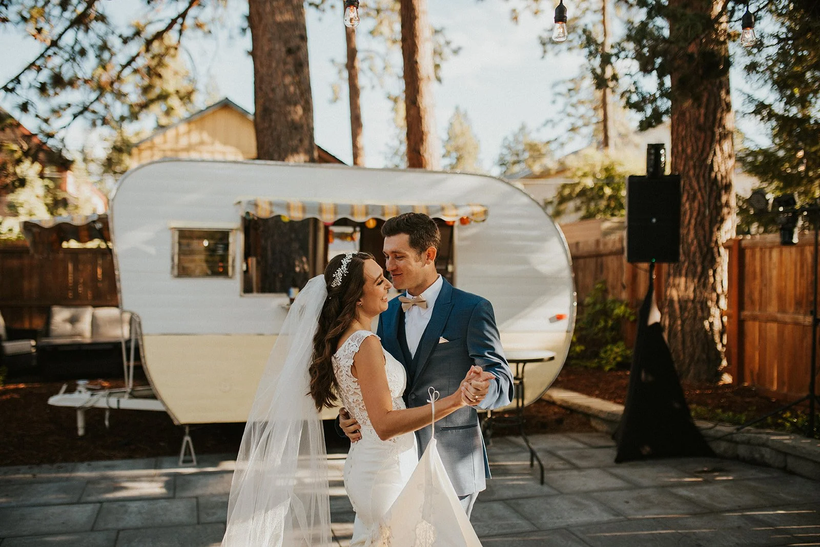 A man and woman dance together in front of a retro trailer in an outdoor courtyard. The woman is in a white wedding dress, and the man is in a blue suit. There are trees in the background against a blue sky.