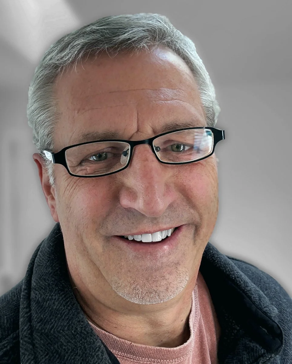 Dave Mensing smiling into the camera. He's wearing glasses, a dark jacket, and set against a soft gray background.