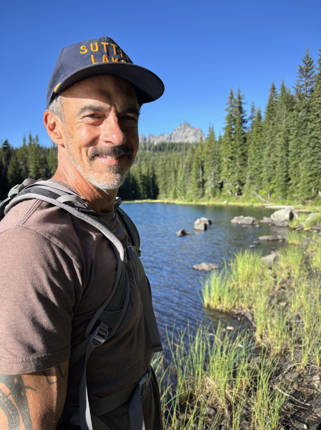 Photo of Dan Rickards smiling at the camera, with a lake, forest, and distant mountain peak in the background