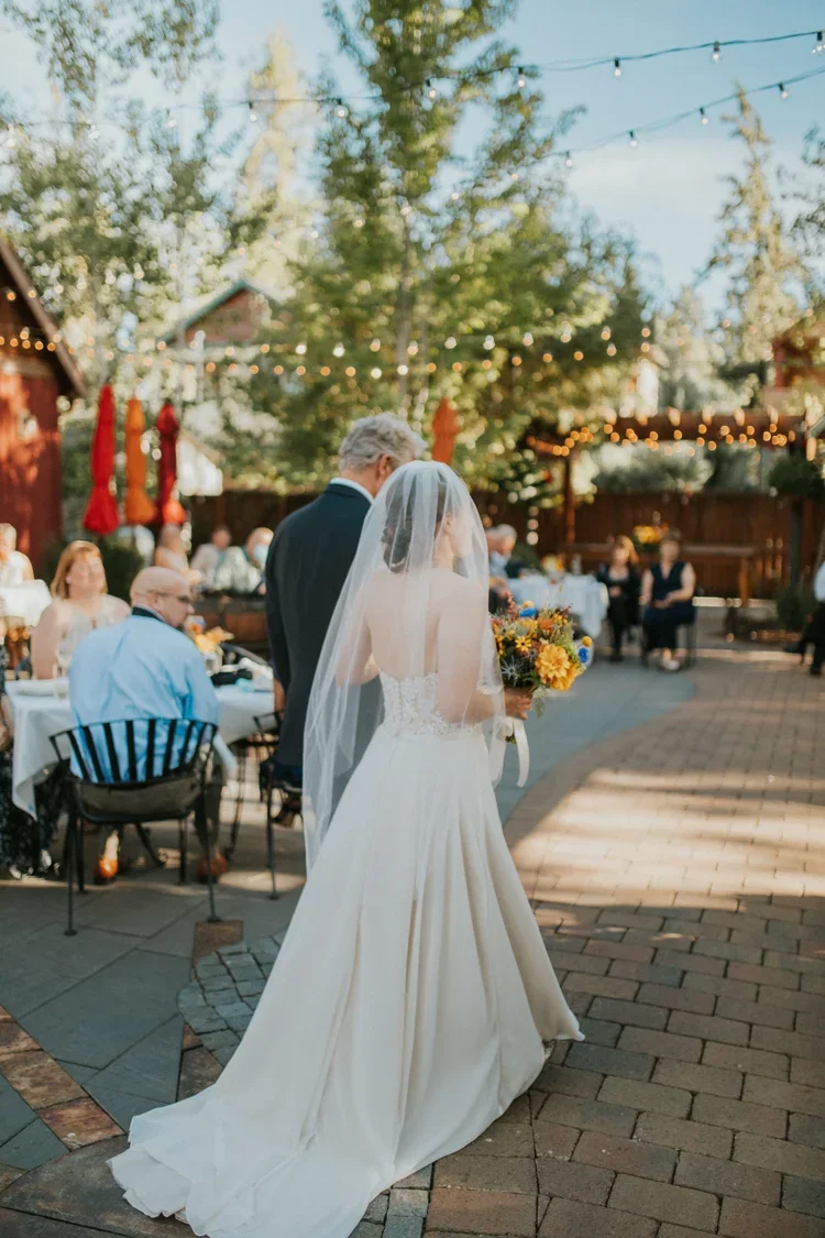 A bride walks down the aisle wearing a white dress while holding the arm of a man in a blue suit in an outdoor wedding venue.