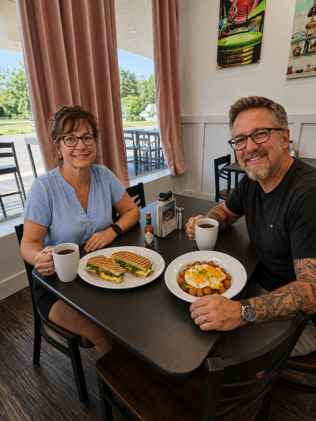 A man and a woman sit at a table in a restaurant, each holding a white coffee mug. The woman has a sandwich on a plate in front of her, and the man has a plate of tater tots topped with melted cheese and a fried egg. The restaurant has pink curtains, framed artwork, and large windows letting in natural light.