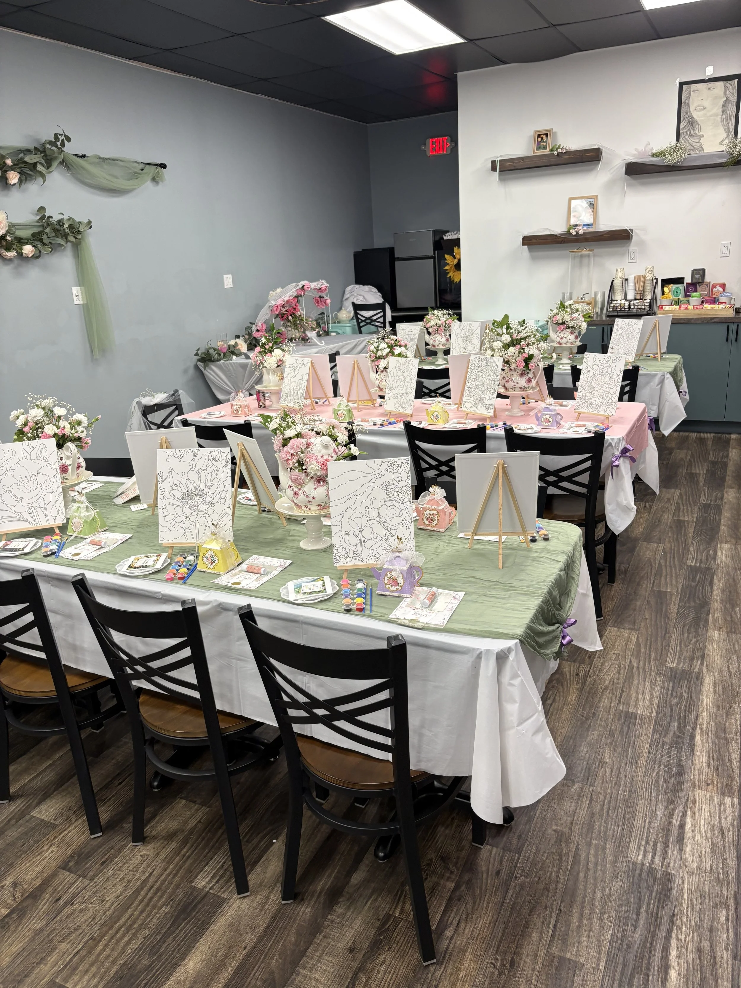 Decorated tables for a crafting or painting activity, with paint palettes, brushes, canvases, floral arrangements, and easels, in a room with light blue and white walls, and shelves with small framed pictures and decorative items.