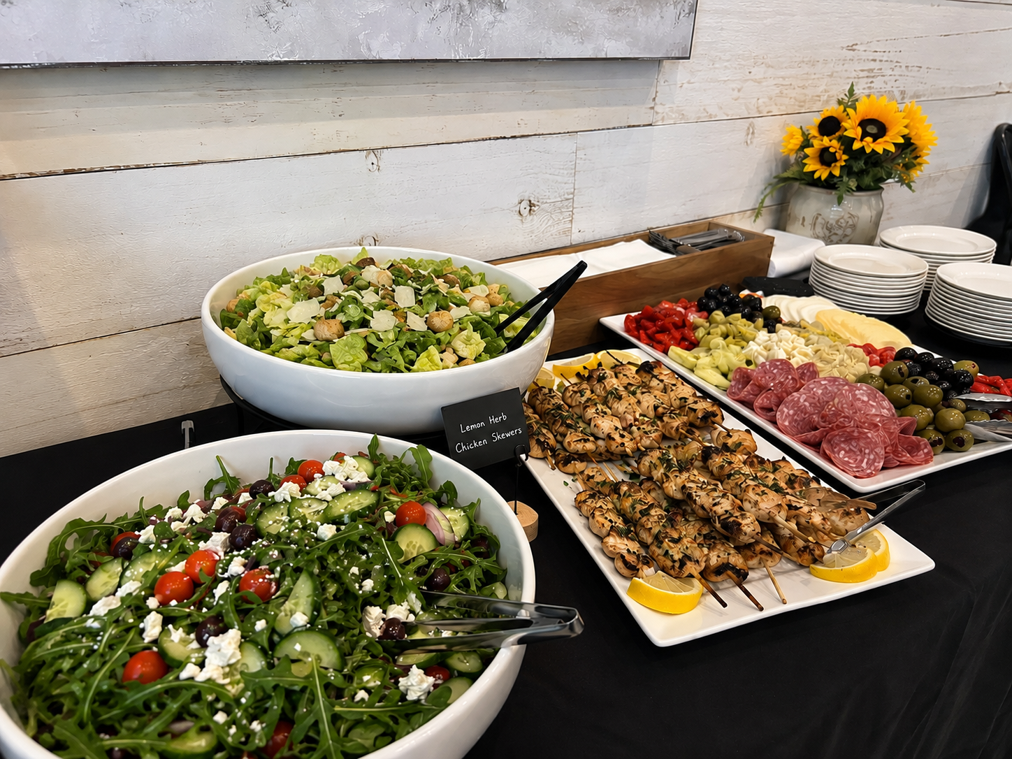 Buffet table with two large salads, grilled chicken skewers, antipasta tray, with a vase of sunflowers in the background.