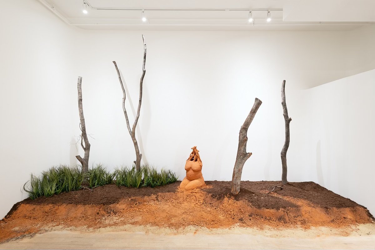 Art installation resembling a small outdoor scene with dirt, four leafless trees, green grass, and a terracotta sculpture as a central focus, displayed in a white-walled gallery.