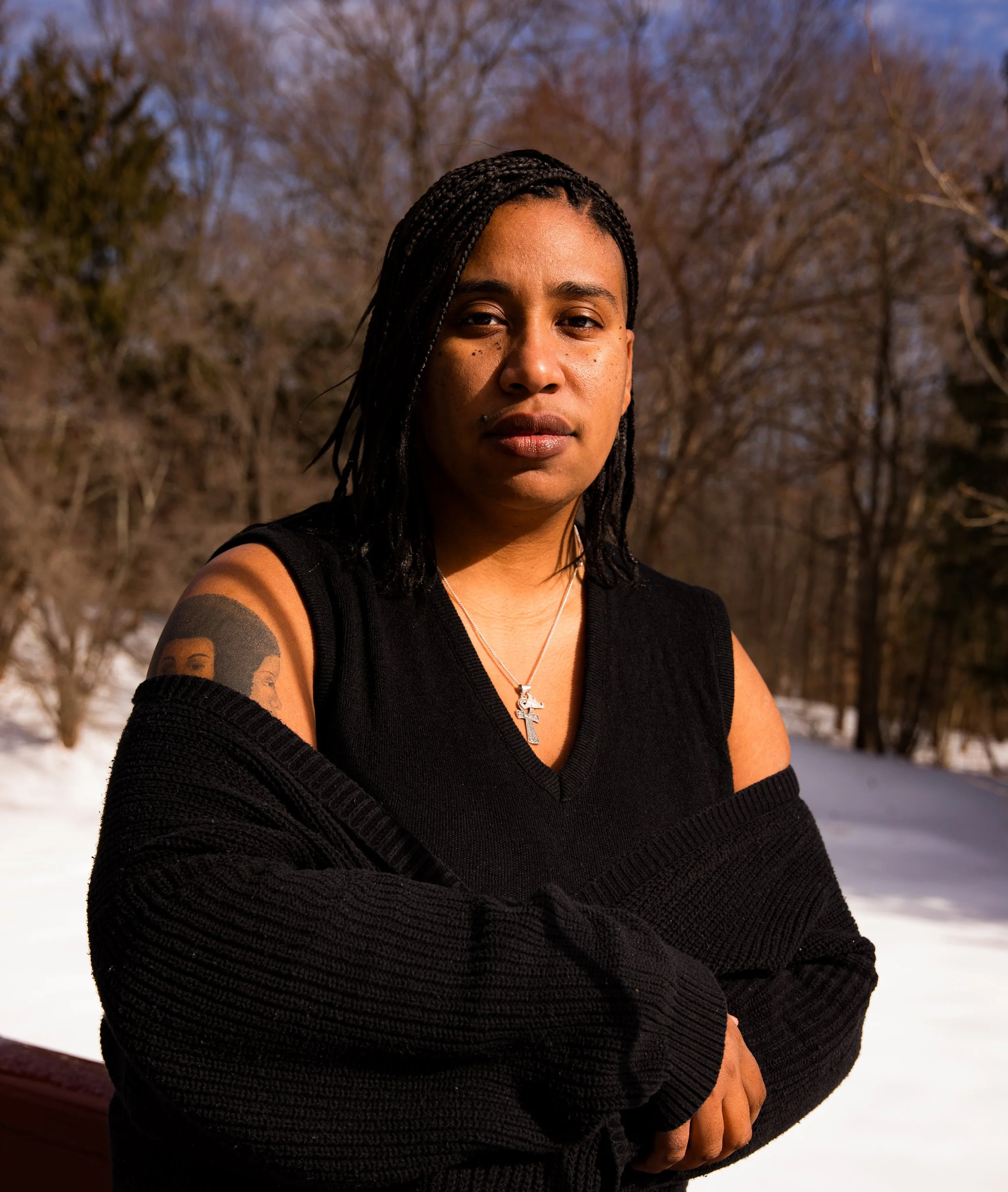 A woman with braided hair wearing a black sleeveless top and a silver cross necklace, standing outdoors in winter with snow on the ground and bare trees in the background.