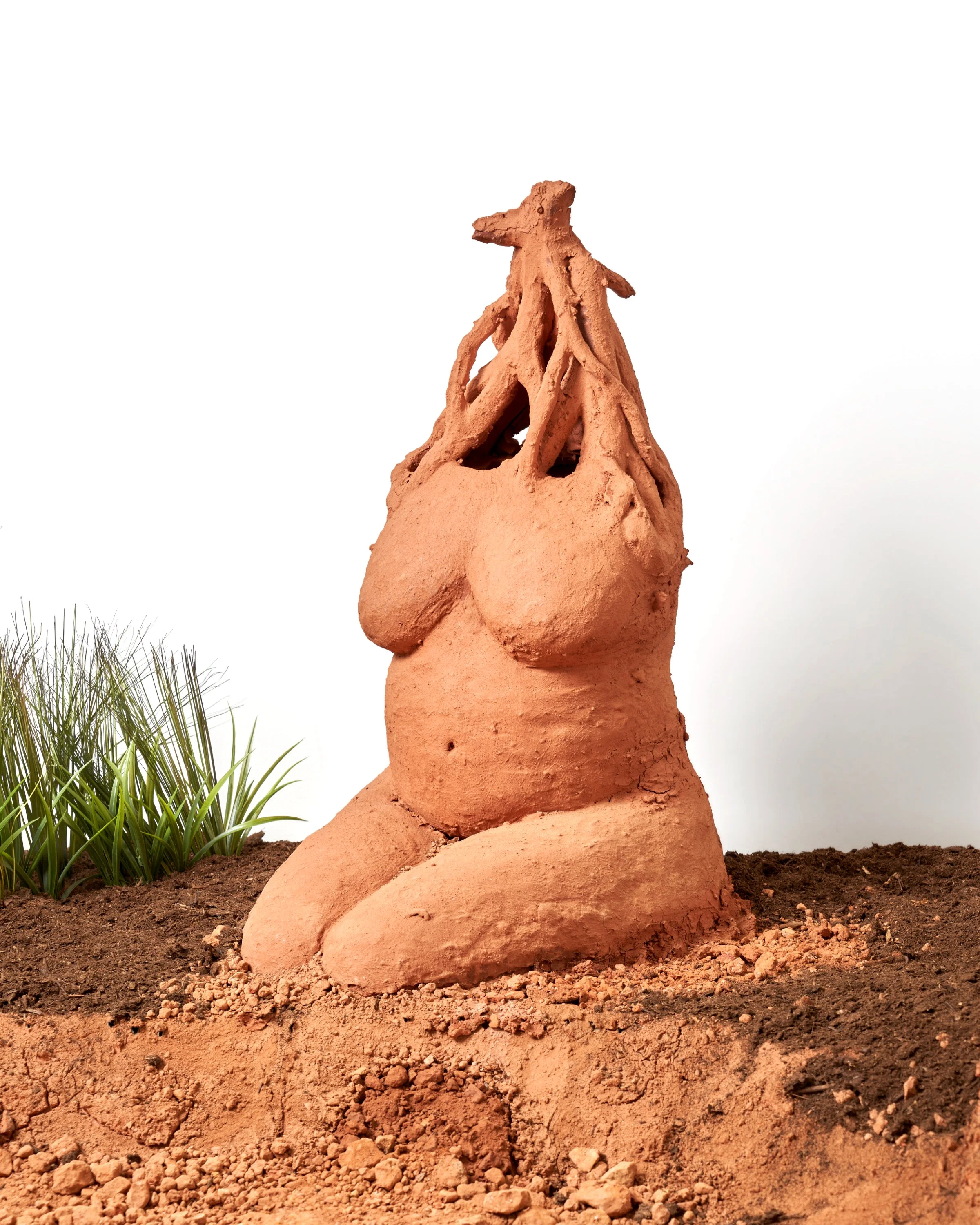 Clay sculpture resembling a humanoid figure with a dragon or dinosaur head, positioned on soil with some grass in the foreground.