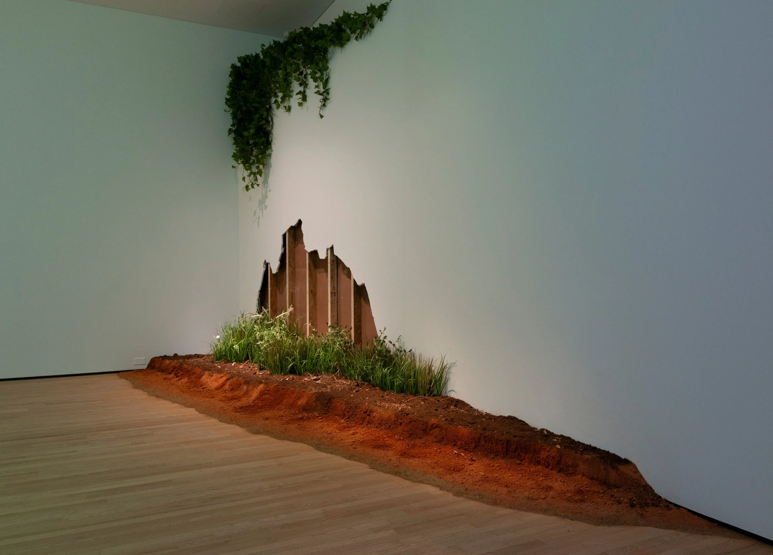 An indoor art installation resembling a diorama with a mound of dirt, green plants, and a cardboard structure of jagged mountain peaks against a plain wall.