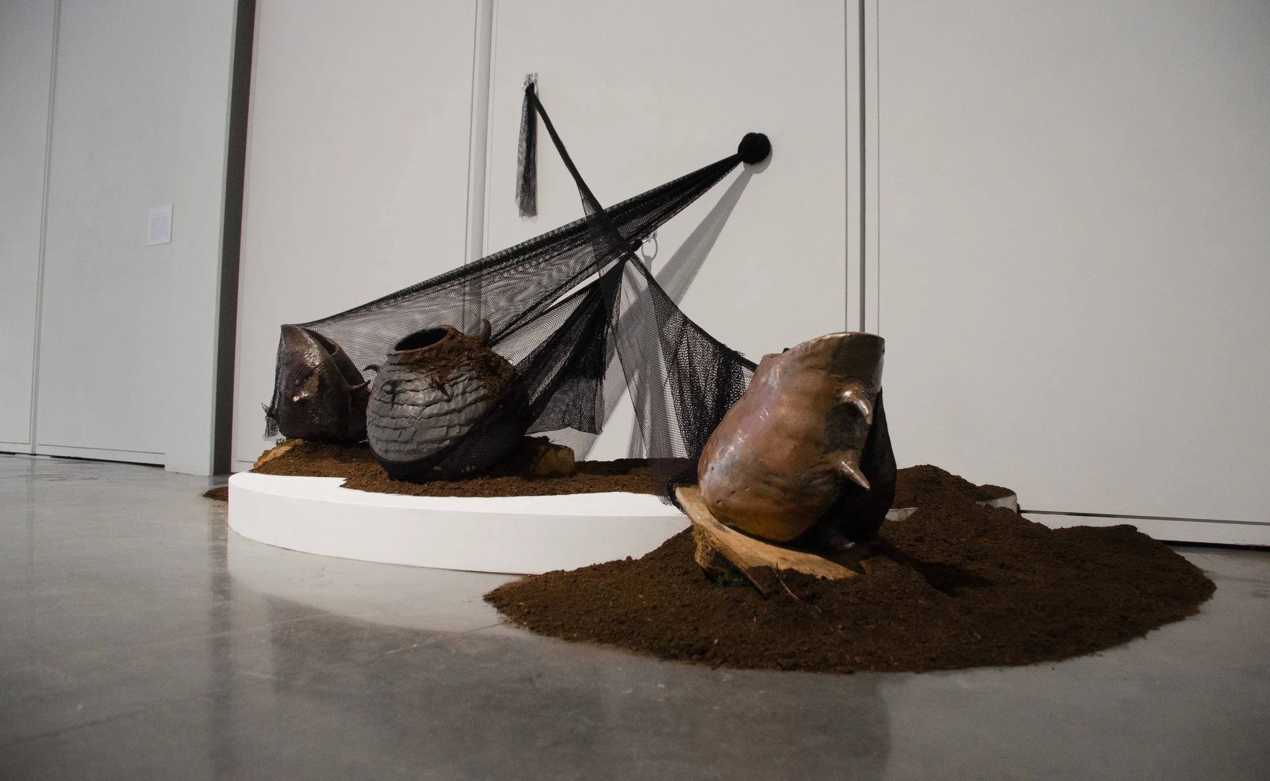 A Collective Struggle
Wooden platform, glazed ceramic, netting, soil, hardware, 2023

Resistance Vessels 1, 2, and 3 are three ceramic vessels constricted
within their own nets that are secured to hardware on the wall. The nets overlap as the
pots pu
