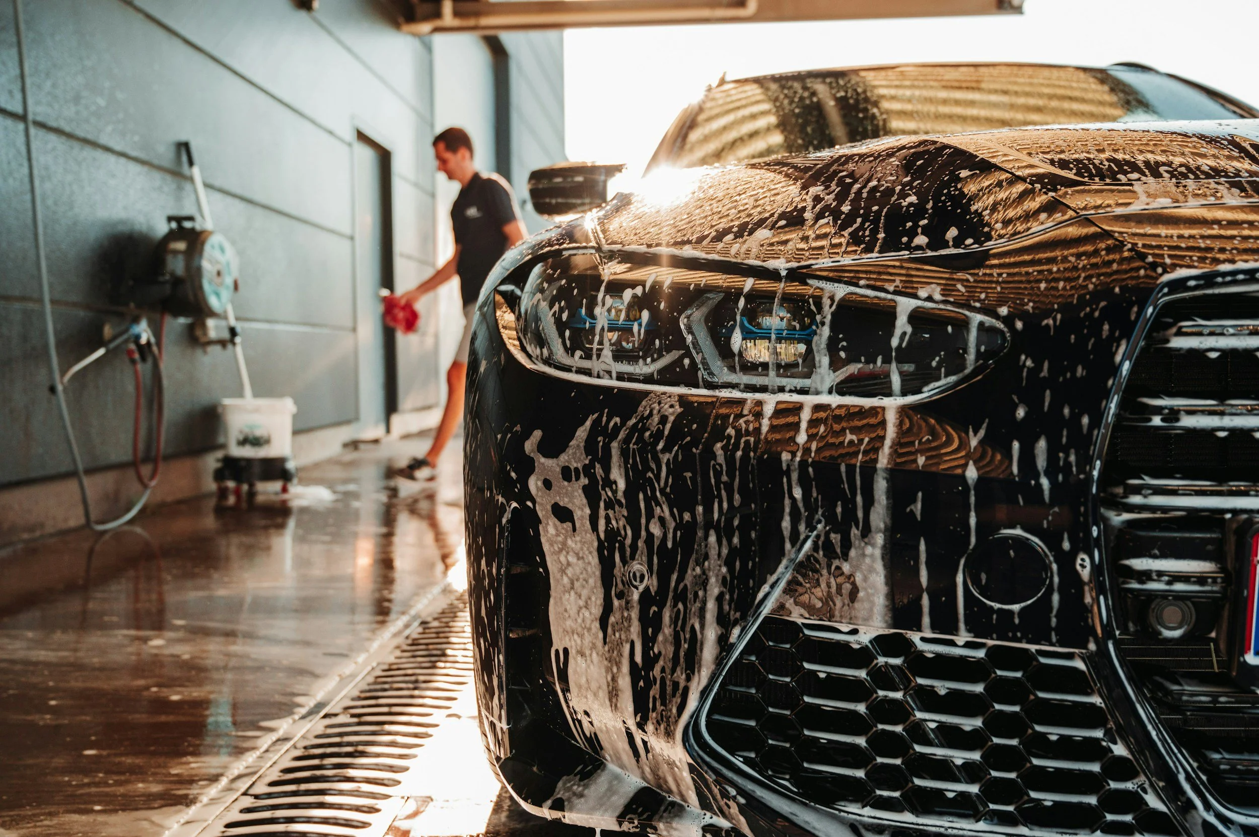 A black car sits in a car wash with soap foam on its front end. A person in the background is cleaning or rinsing a window or door, with sunlight shining behind them.