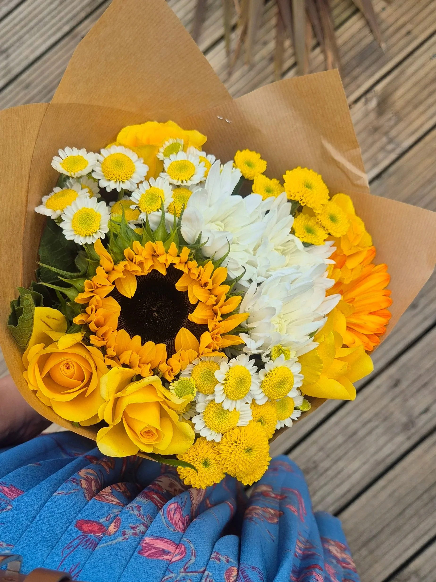 A colorful bouquet of yellow, white, and orange flowers held by a person wearing a blue dress with floral patterns, standing outdoors on a wooden surface.