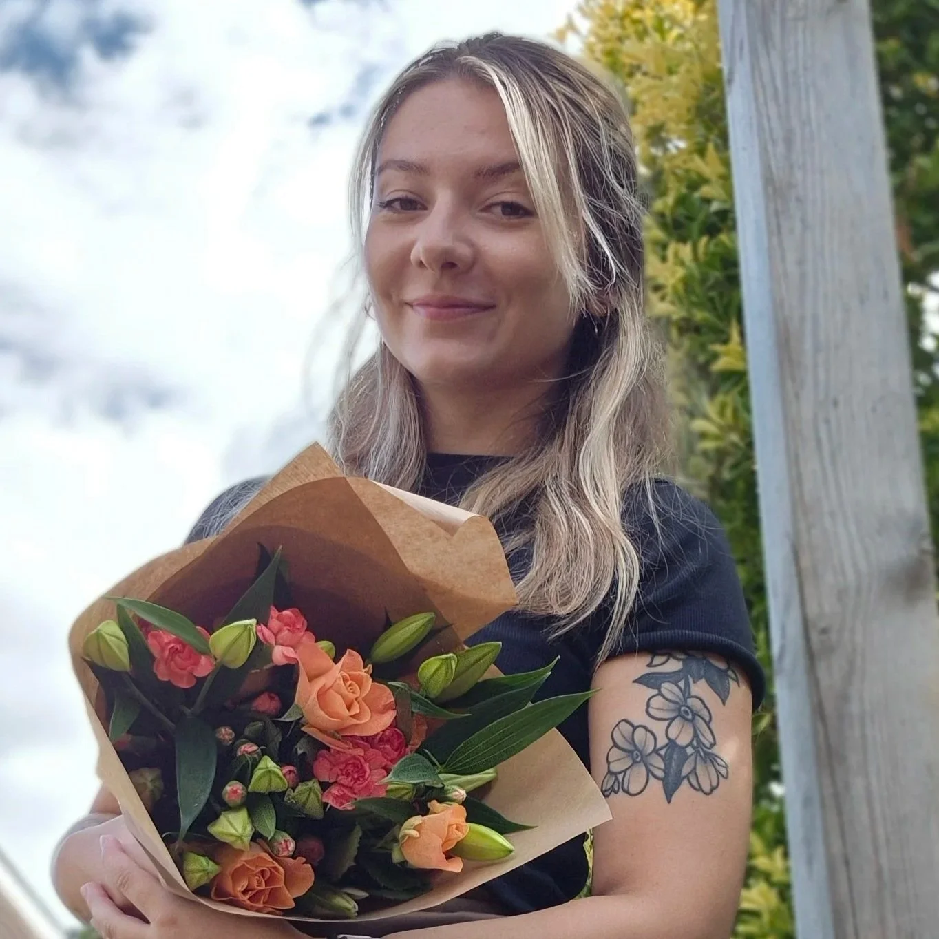 A young woman with blonde hair holding a bouquet of flowers wrapped in brown paper, standing outdoors near a tree with green leaves in the background.