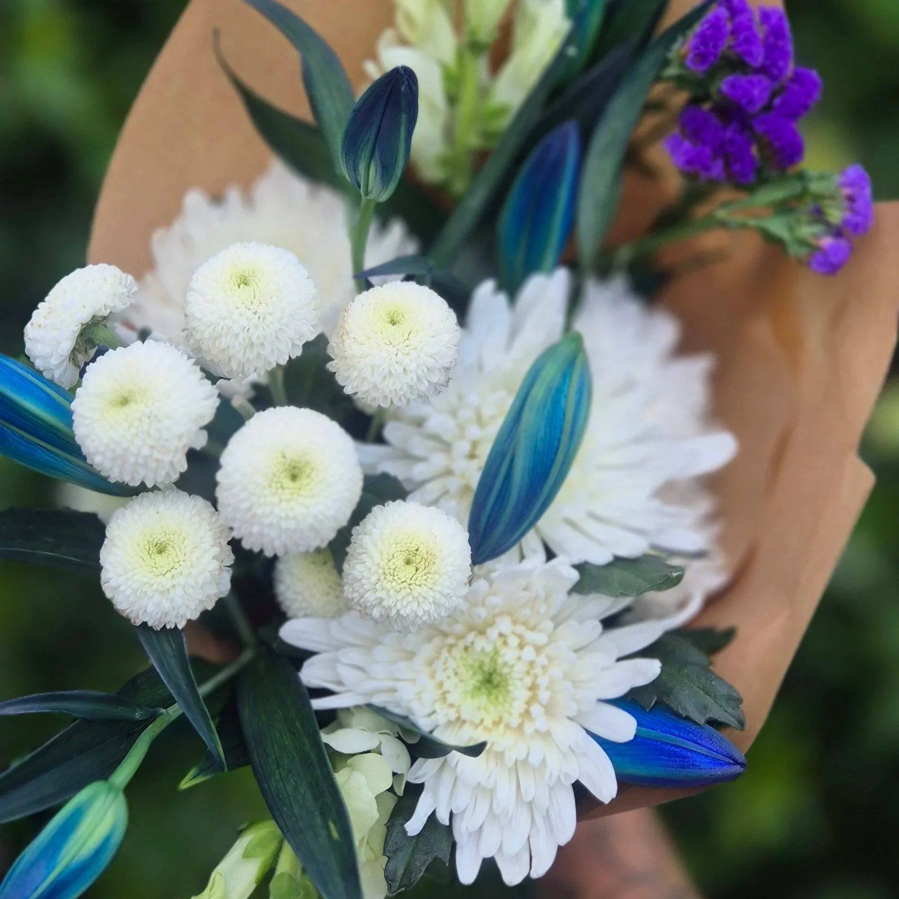 A bouquet of white and purple flowers with green leaves, wrapped in brown paper.