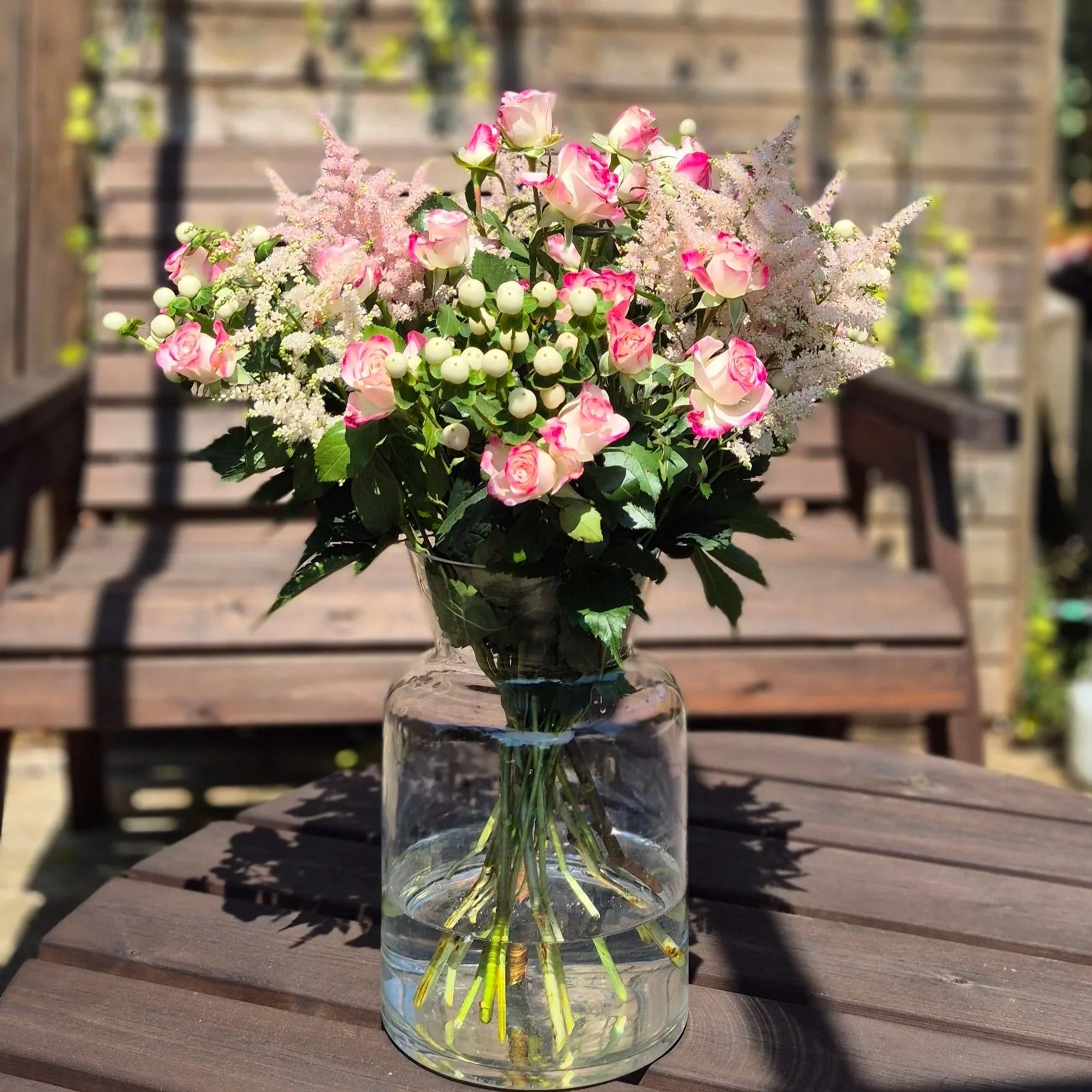 A glass vase with a bouquet of pink and white roses, pink astilbe, and white berries, placed on a wooden table outdoors with sunlight and blurred wooden background.