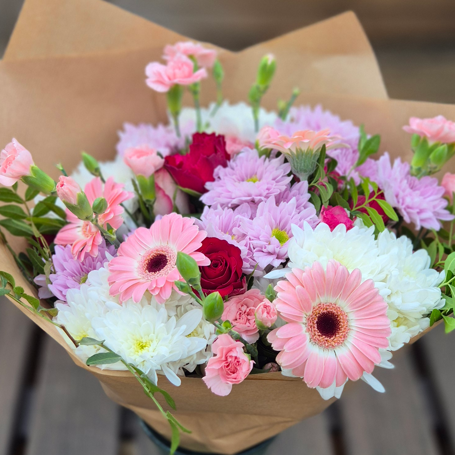 A large bouquet of pink, white, and red flowers wrapped in brown paper. Featuring roses, spray carnations to create a decadent and feminine feel.