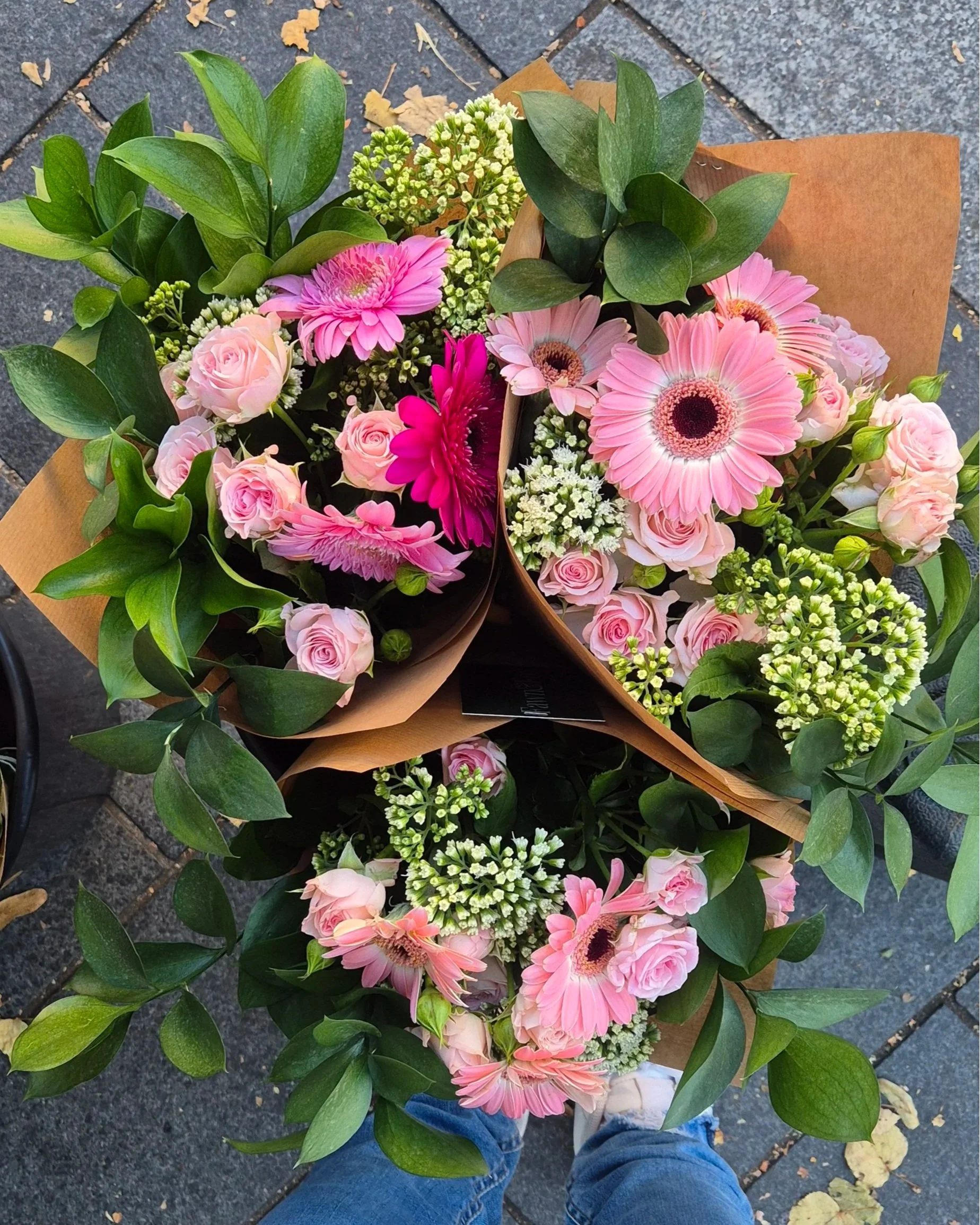 Top-down view of a bouquet of pink, white, and green flowers and leaves on a sidewalk with a person's feet visible in jeans and shoes.