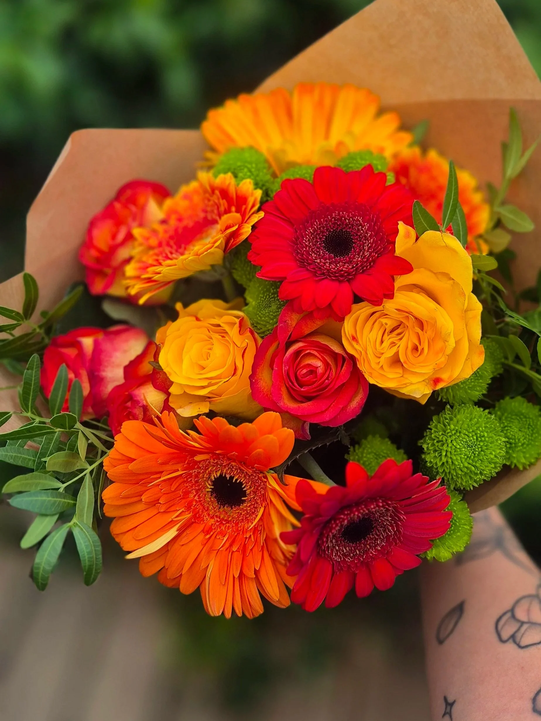 A colorful bouquet of flowers including red, orange, yellow, and green flowers held by a person with tattoos on their arm.