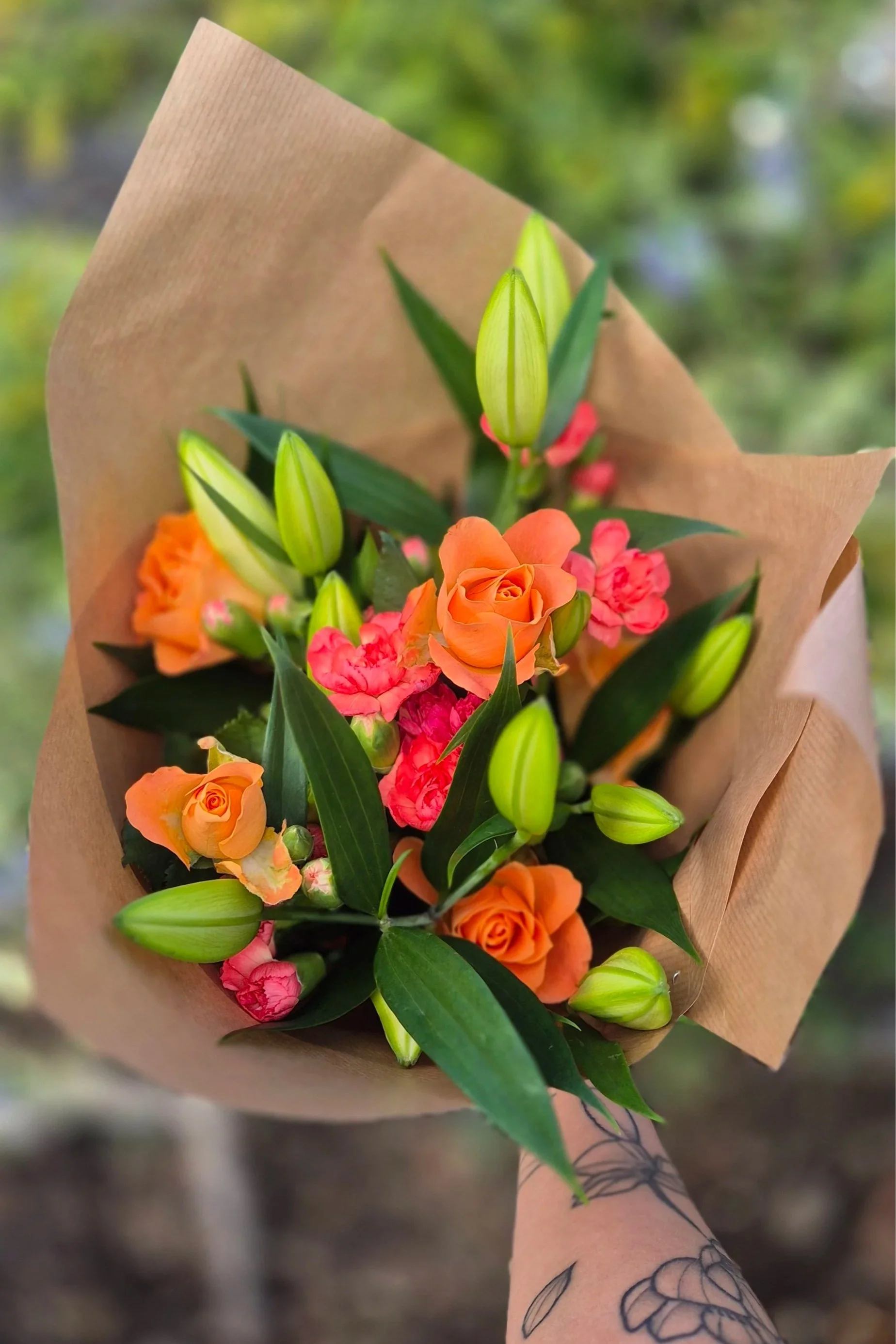 Hand holding a flower bouquet wrapped in brown paper, containing orange roses, pink flowers, and green lilies with closed buds, outdoors with blurred green background.
