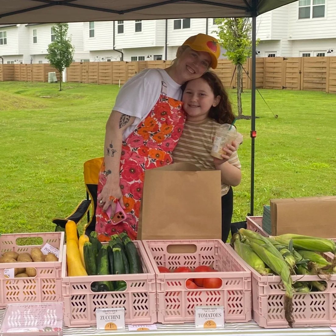 Smiling girl standing next to woman in colorful apron at a farmer's market stall selling fresh vegetables, including zucchini, tomatoes, and corn, under a canopy with a grassy yard and houses in the background.