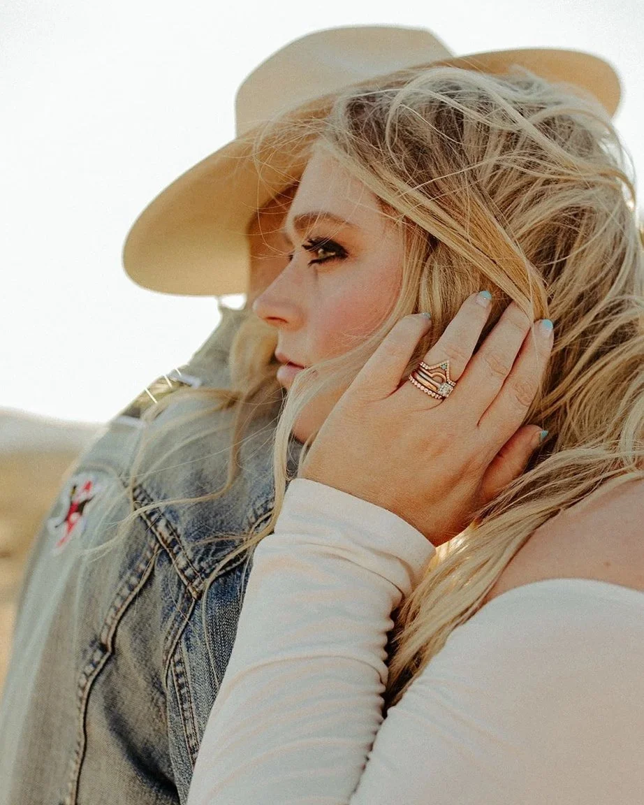 Close-up of a blonde woman touching her hair, wearing rings and a white top, with a person in a cowboy hat in the background.