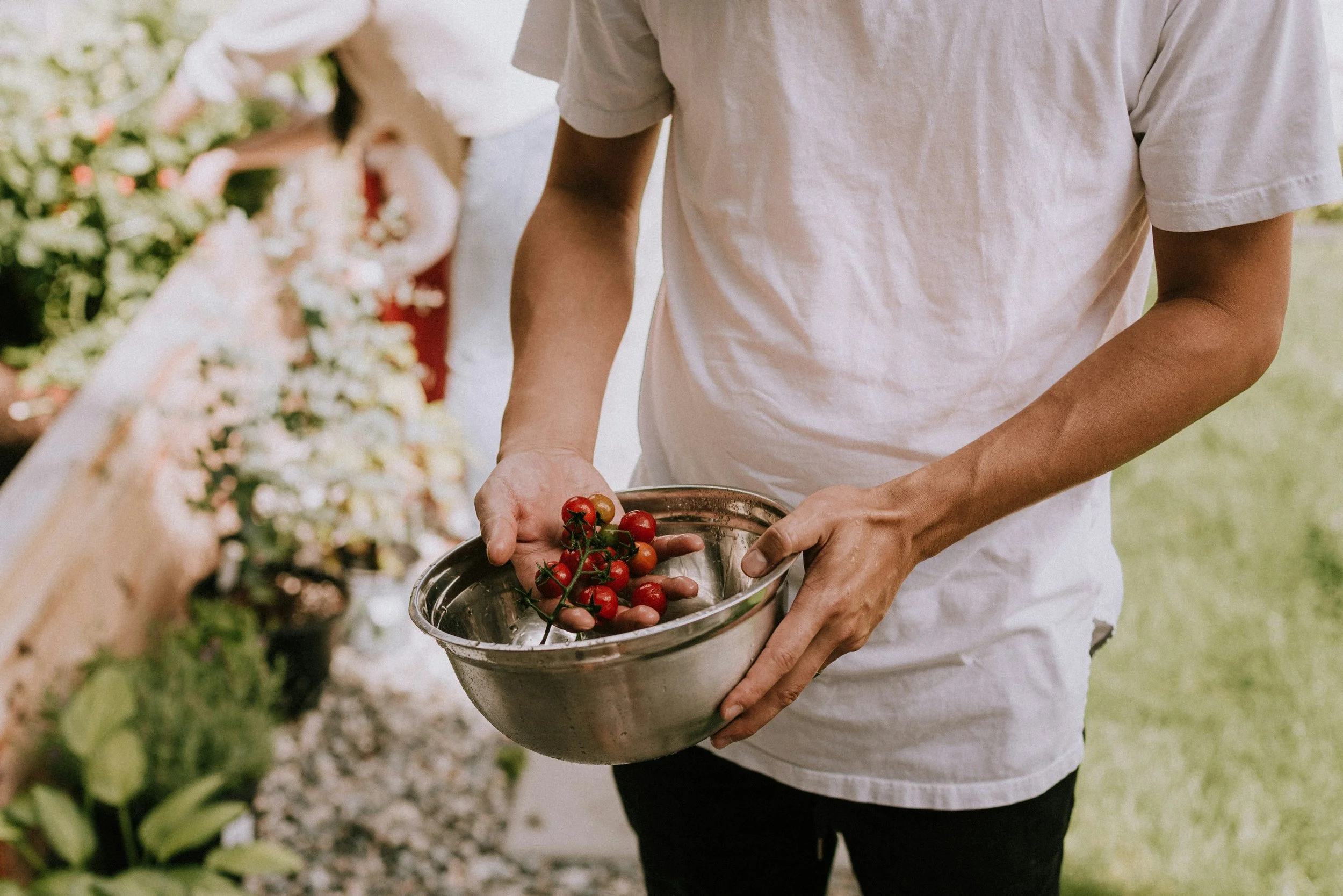 Person holding a metal bowl of cherry tomatoes at an outdoor event with floral decorations in the background.