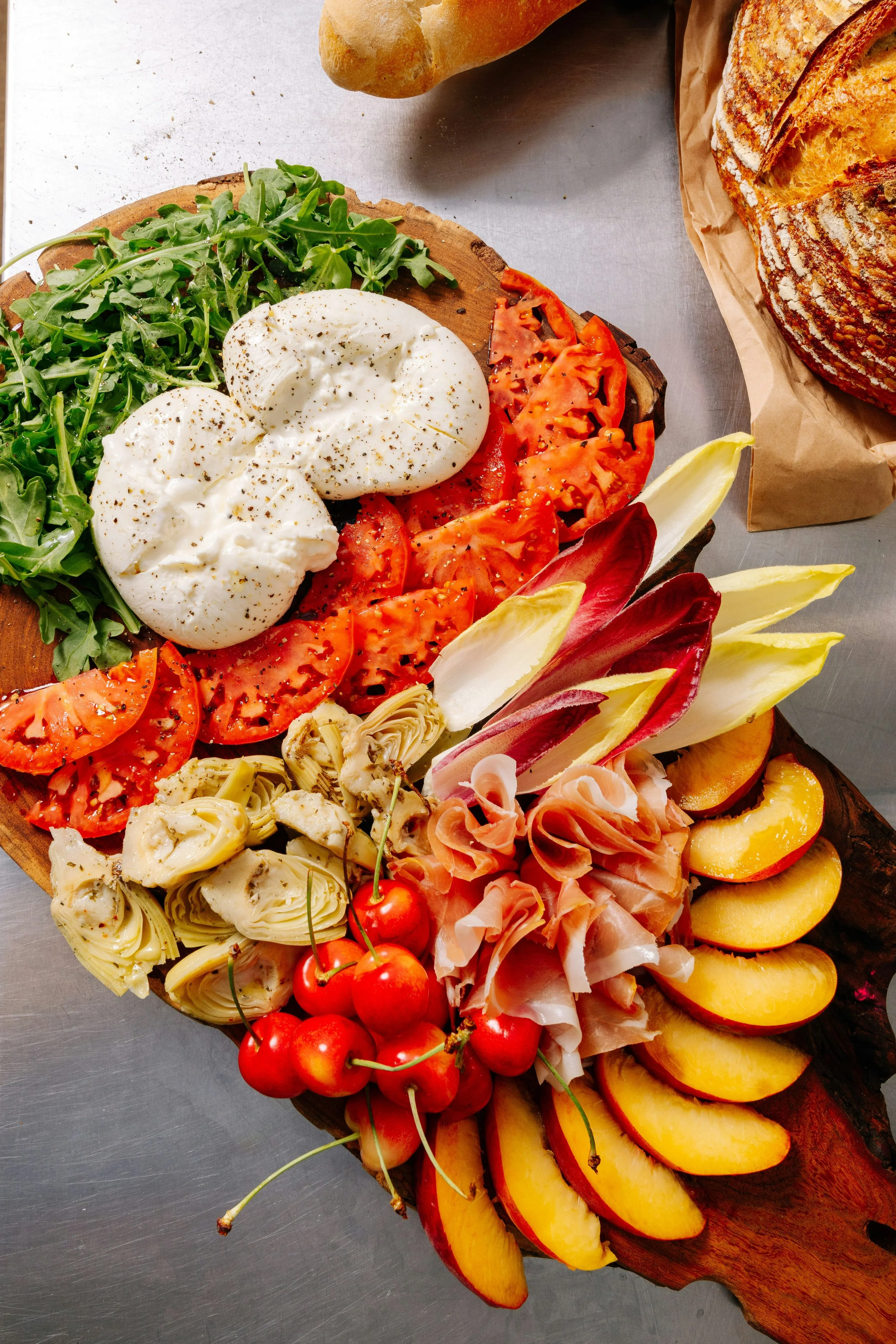 A wooden platter with a fresh salad including arugula, sliced tomatoes, mozzarella cheese, artichoke hearts, and cherries. Sliced bread and a baguette are visible in the background.