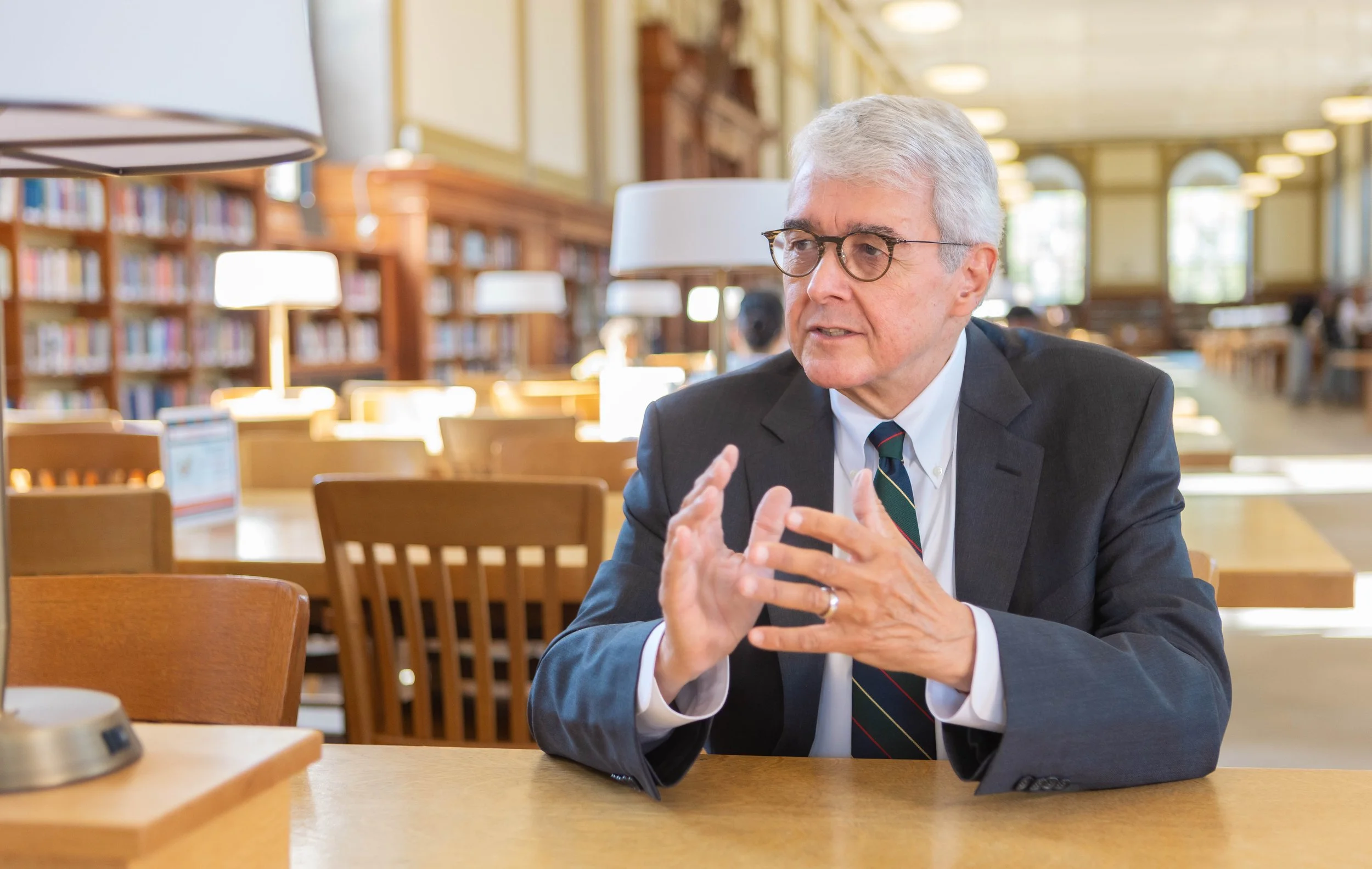 An older man with gray hair and glasses wearing a dark suit, white shirt, and striped tie, sitting at a wooden table in a library, engaged in conversation or explaining something, with shelves of books and bright lamps in the background.