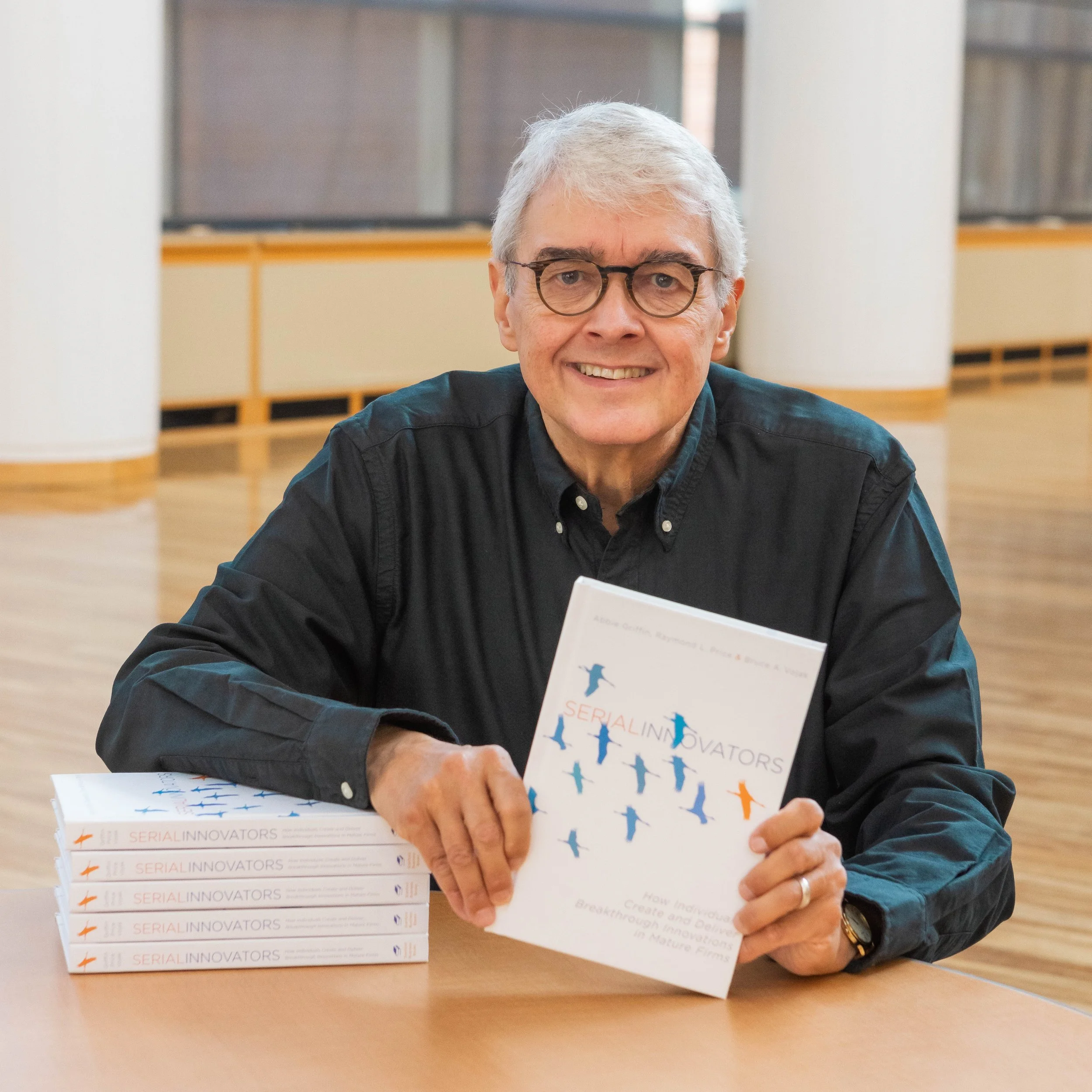 An older man with gray hair and glasses, smiling, sitting at a table with several copies of a book titled 'Serial Innovators.' The background is an indoor space with windows and a wooden structure.