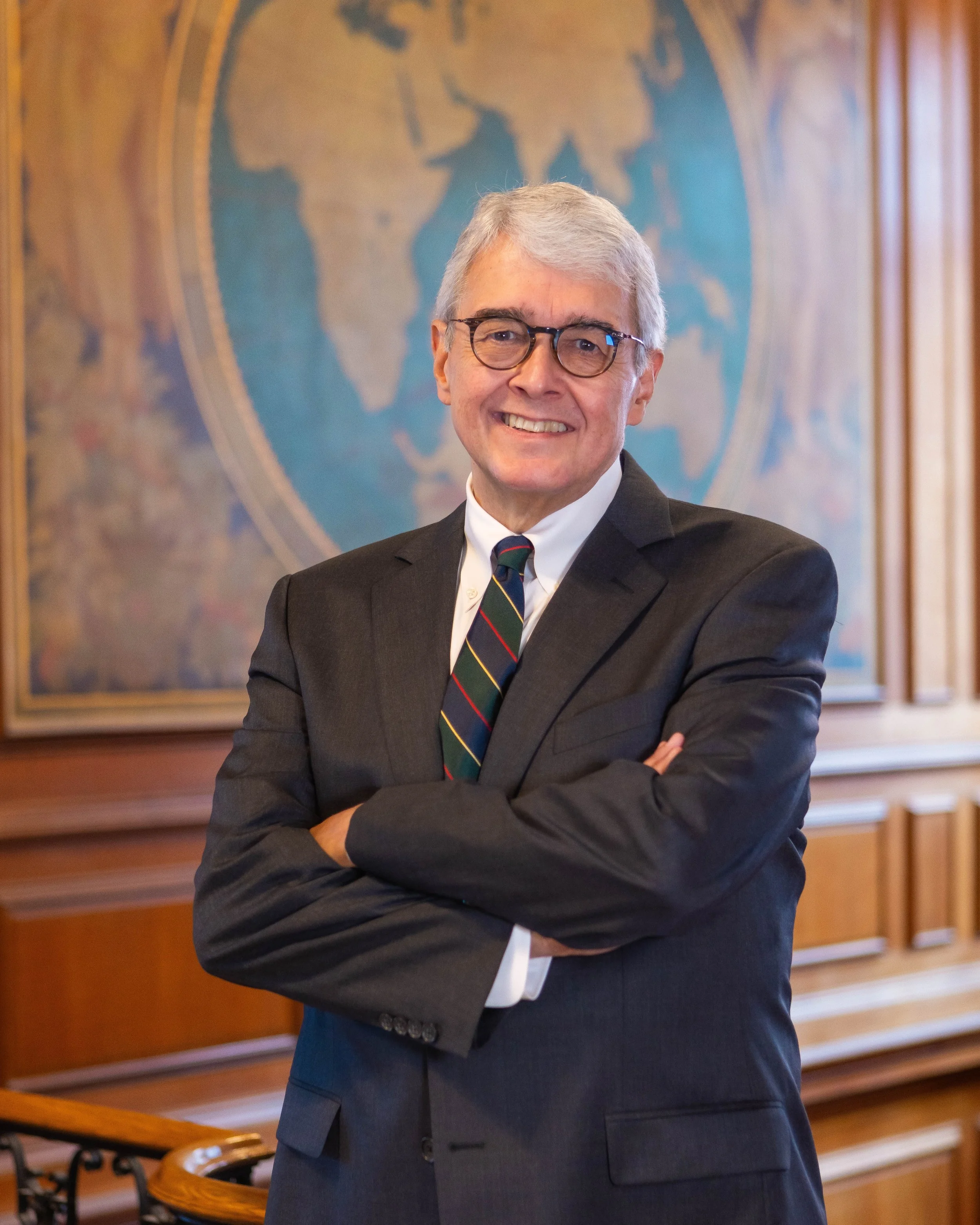 A smiling man with gray hair and glasses, dressed in a dark suit and a striped tie, standing with crossed arms in front of a wall with a world map poster.
