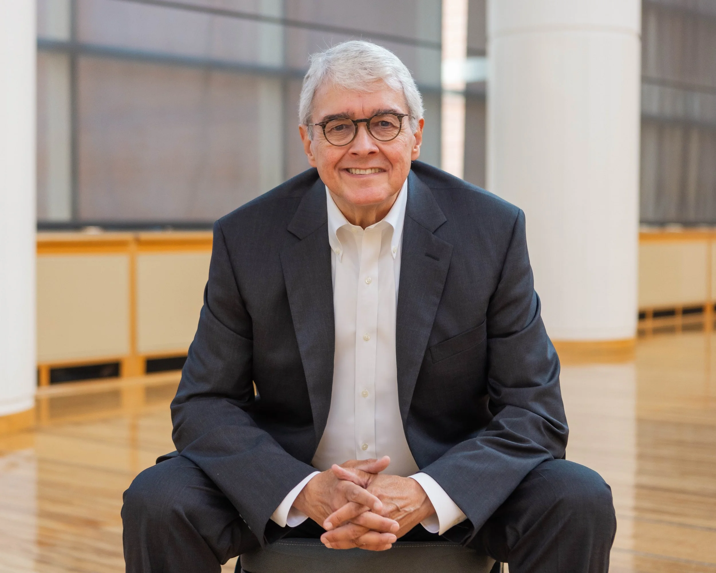 A smiling elderly man with gray hair and glasses, wearing a dark suit and white shirt, sitting on the floor with hands clasped in a modern, well-lit building with large windows and wooden accents.