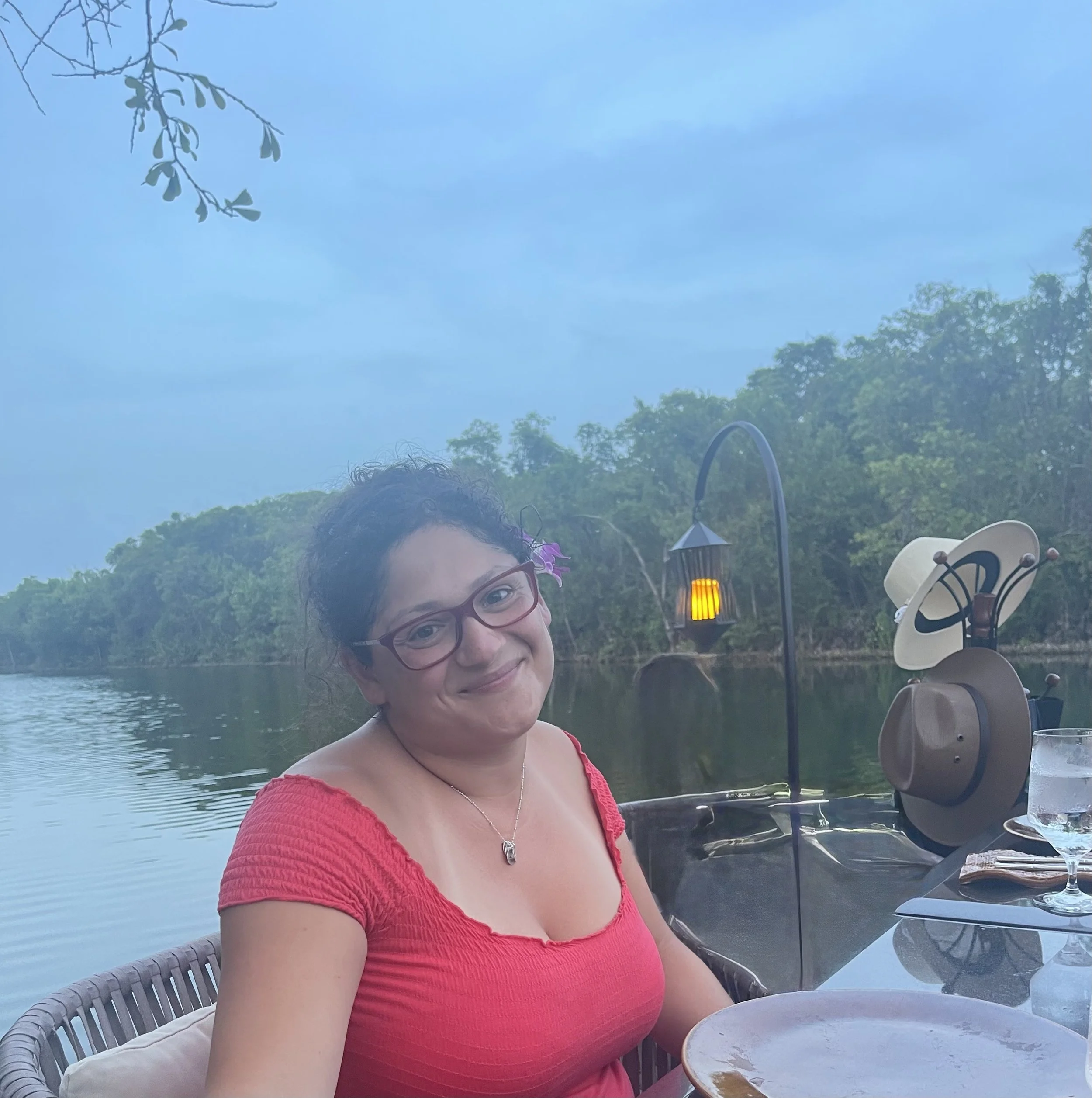 A smiling woman with glasses and curly hair wearing a red top sitting at an outdoor table near a lake, with trees and a blue sky in the background.