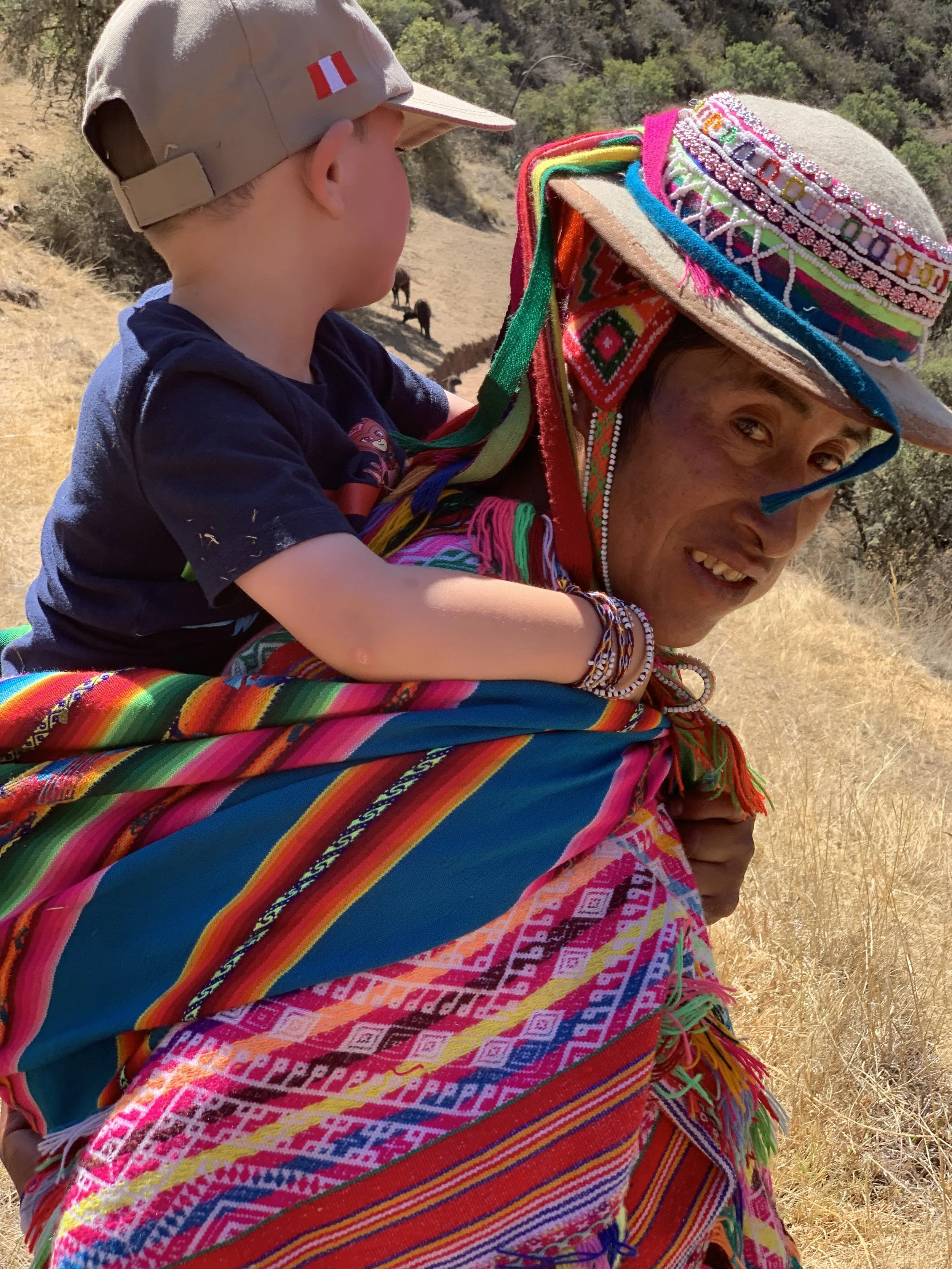 A person dressed in traditional, colorful Peruvian clothing carrying a young boy on their back, outdoors in a dry, grassy area with trees in the background.