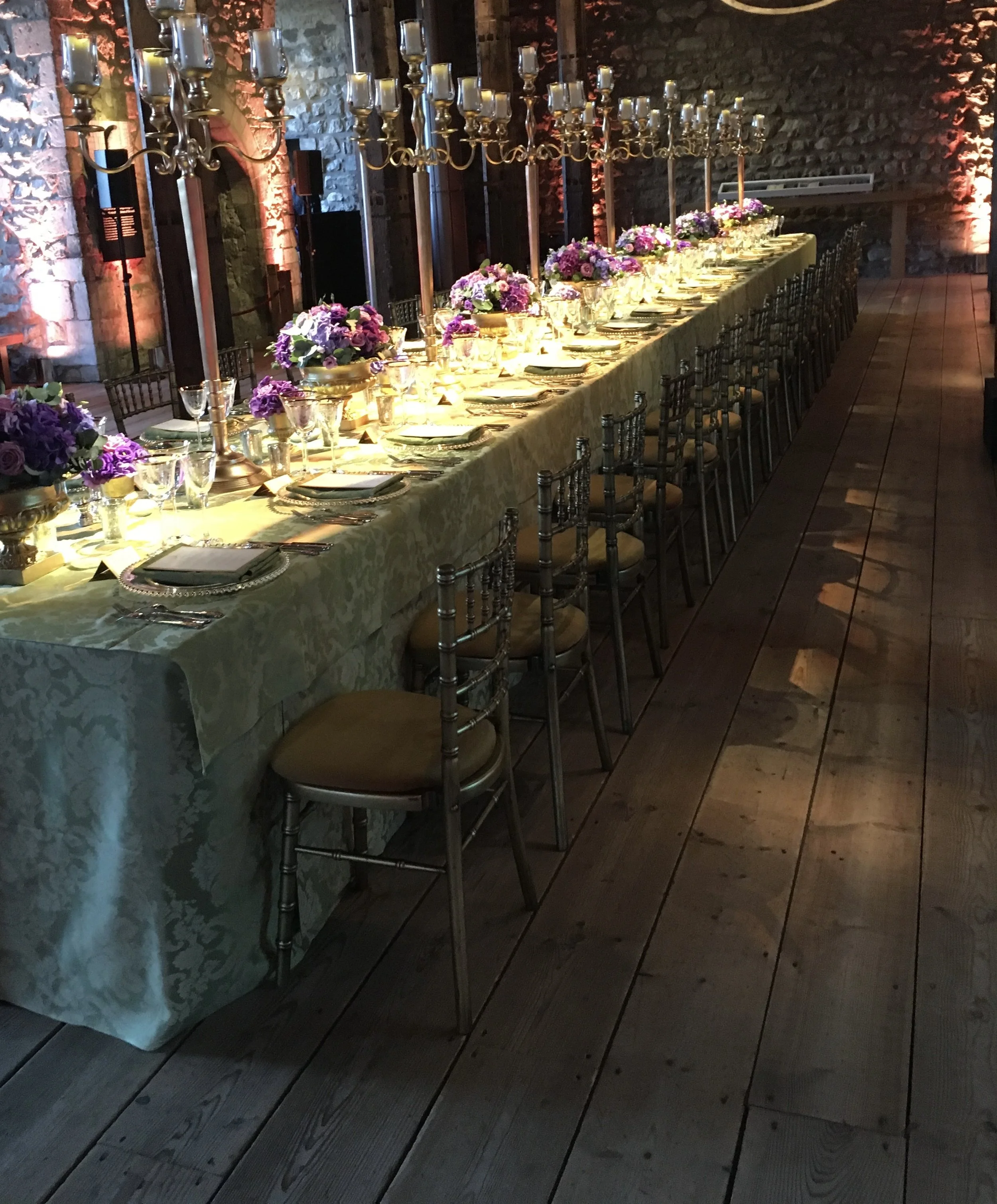 A long rectangular table set for a formal dinner against a brick wall with colored uplighting. The table is covered with a yellow tablecloth and decorated with multiple flower arrangements and tall candelabras. Each place setting includes a plate, silverware, a napkin, and glasses.