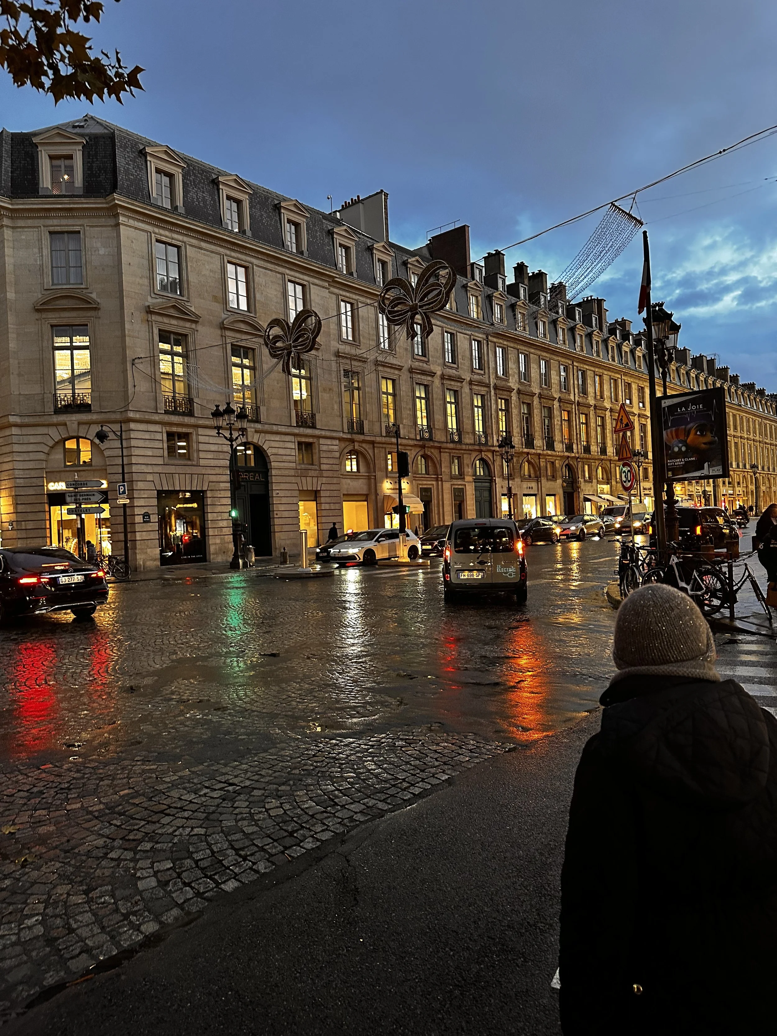 A street scene in the evening with historic Parisian-style buildings illuminated by warm lights, wet cobblestone street reflecting the lights, parked cars, bicycles, and decorative bows hanging above the street, with a person in dark clothing and a knit hat in the foreground.