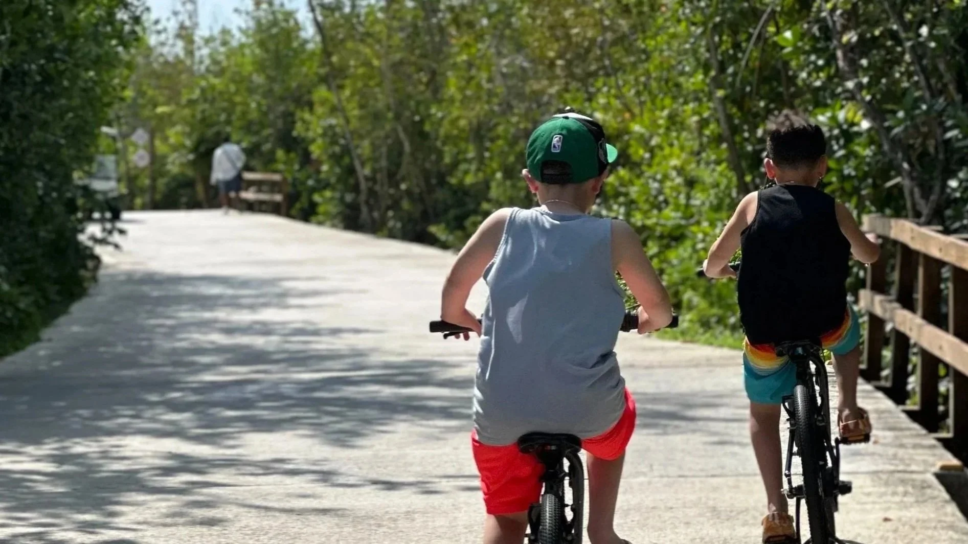 Two children riding bikes on a sunny, tree-lined path with a wooden railing, one wearing a green cap and sleeveless shirt, the other in a black tank top with rainbow shorts.