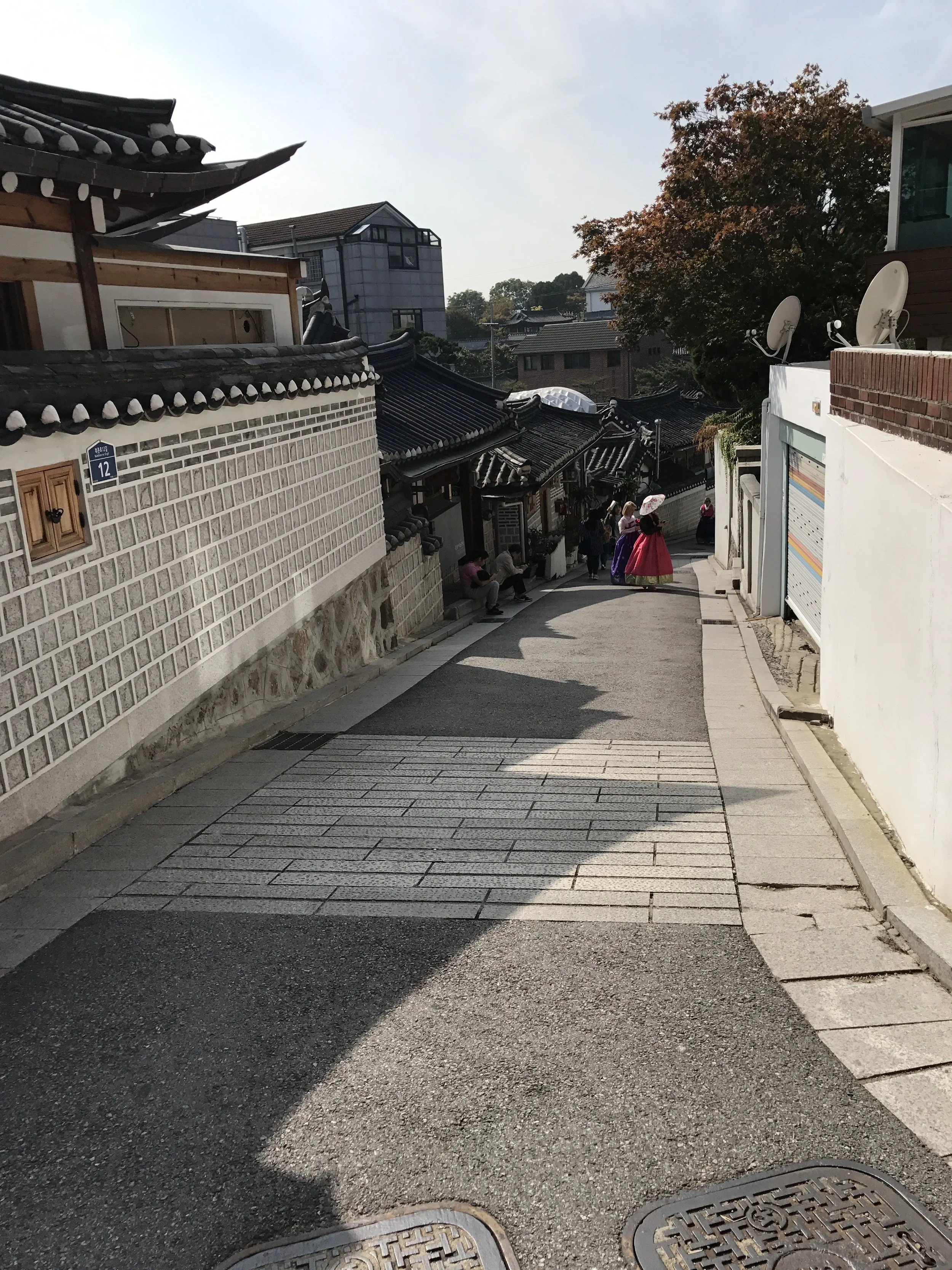 A street with traditional Korean-style houses, some modern buildings, and a group of people walking, including a woman in a pink hanbok holding an umbrella, on a sunny day with clear skies.