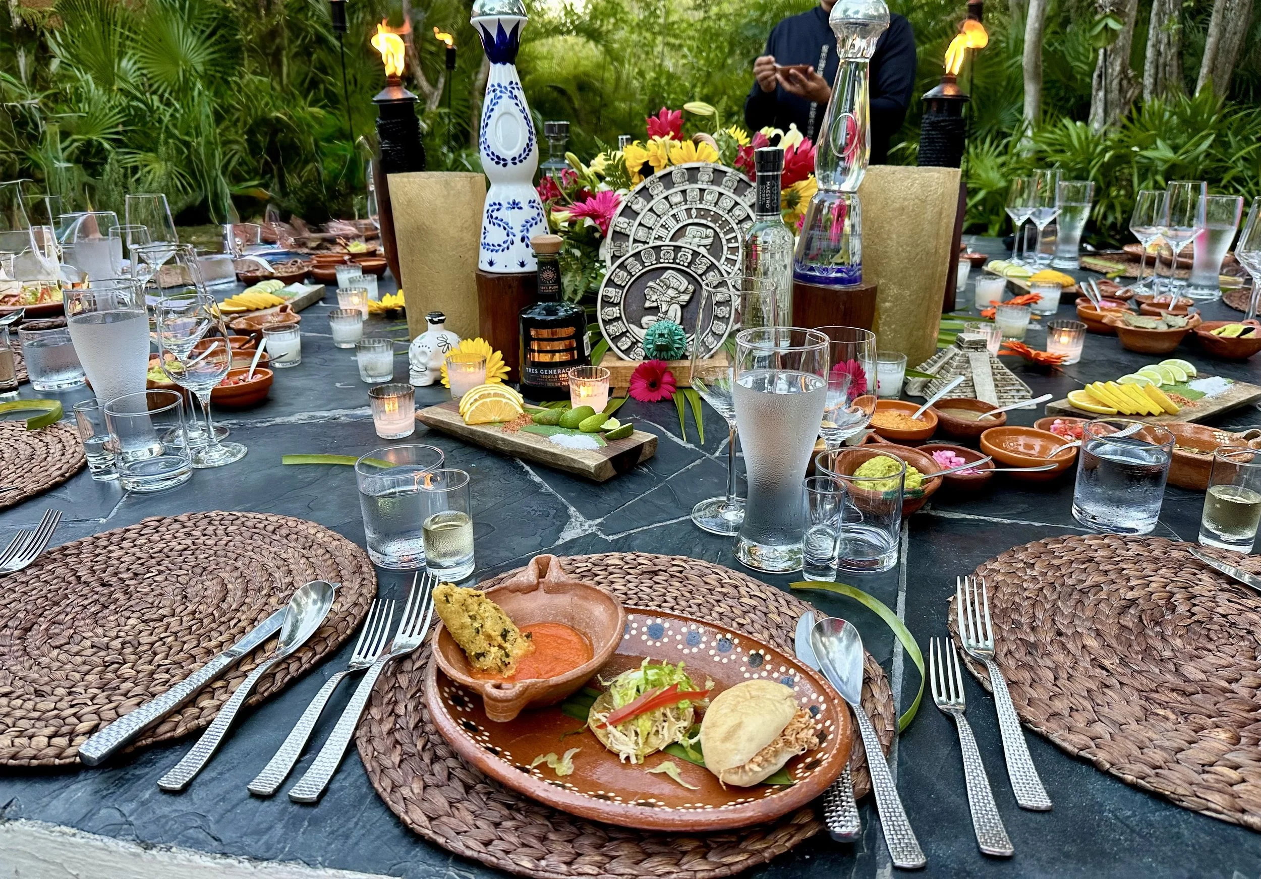 A festive outdoor dining setup with a large round table covered in various Mexican dishes, including tacos, salsa, and lemon slices. The table has woven placemats and silverware, surrounded by glasses of water. In the center, there are colorful flowers, vases, candles, and decorative items, with greenery in the background.