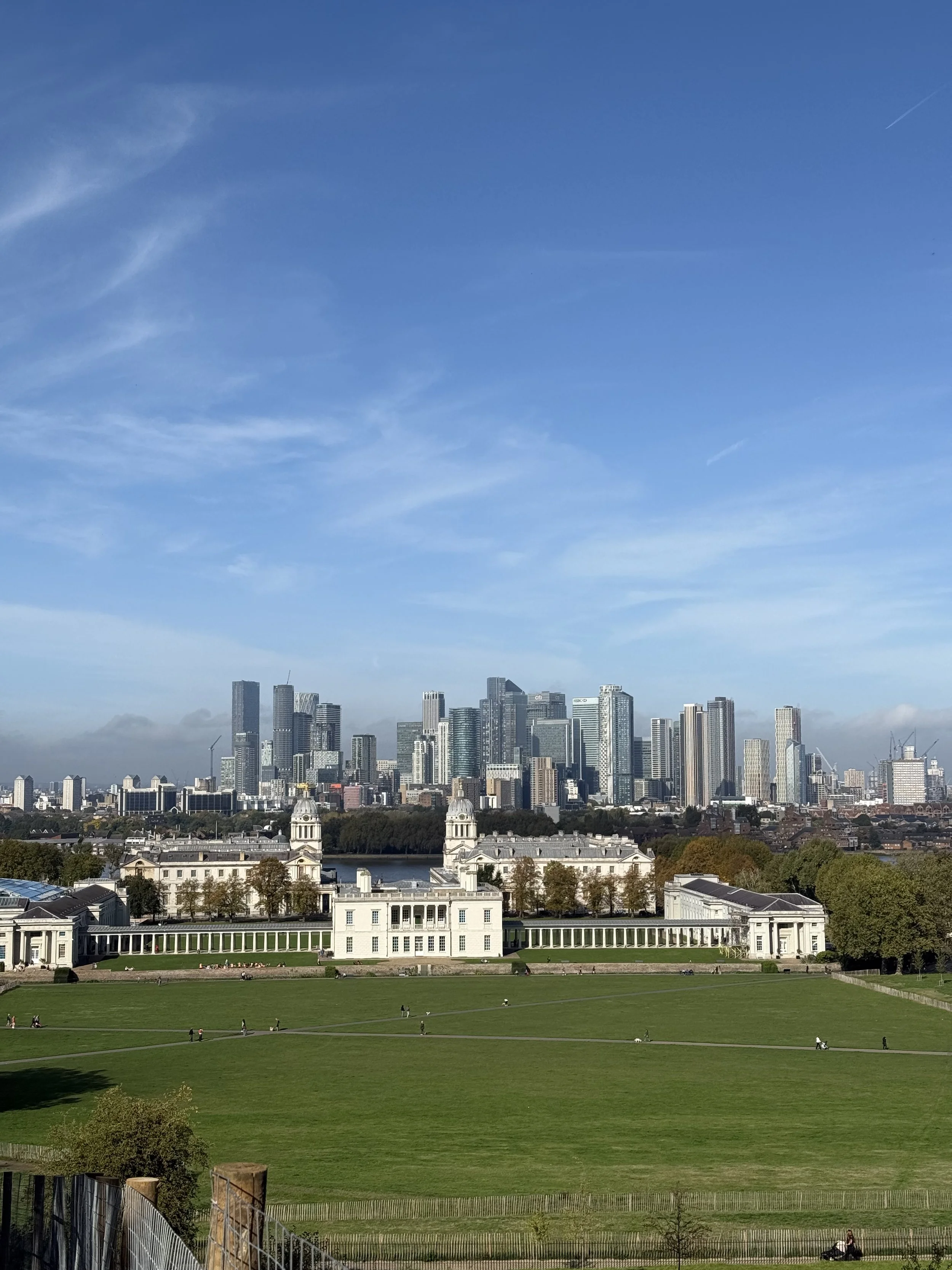A city skyline with tall skyscrapers in the background, a historic white mansion in the midground, green fields with people walking or relaxing, and a clear blue sky above.