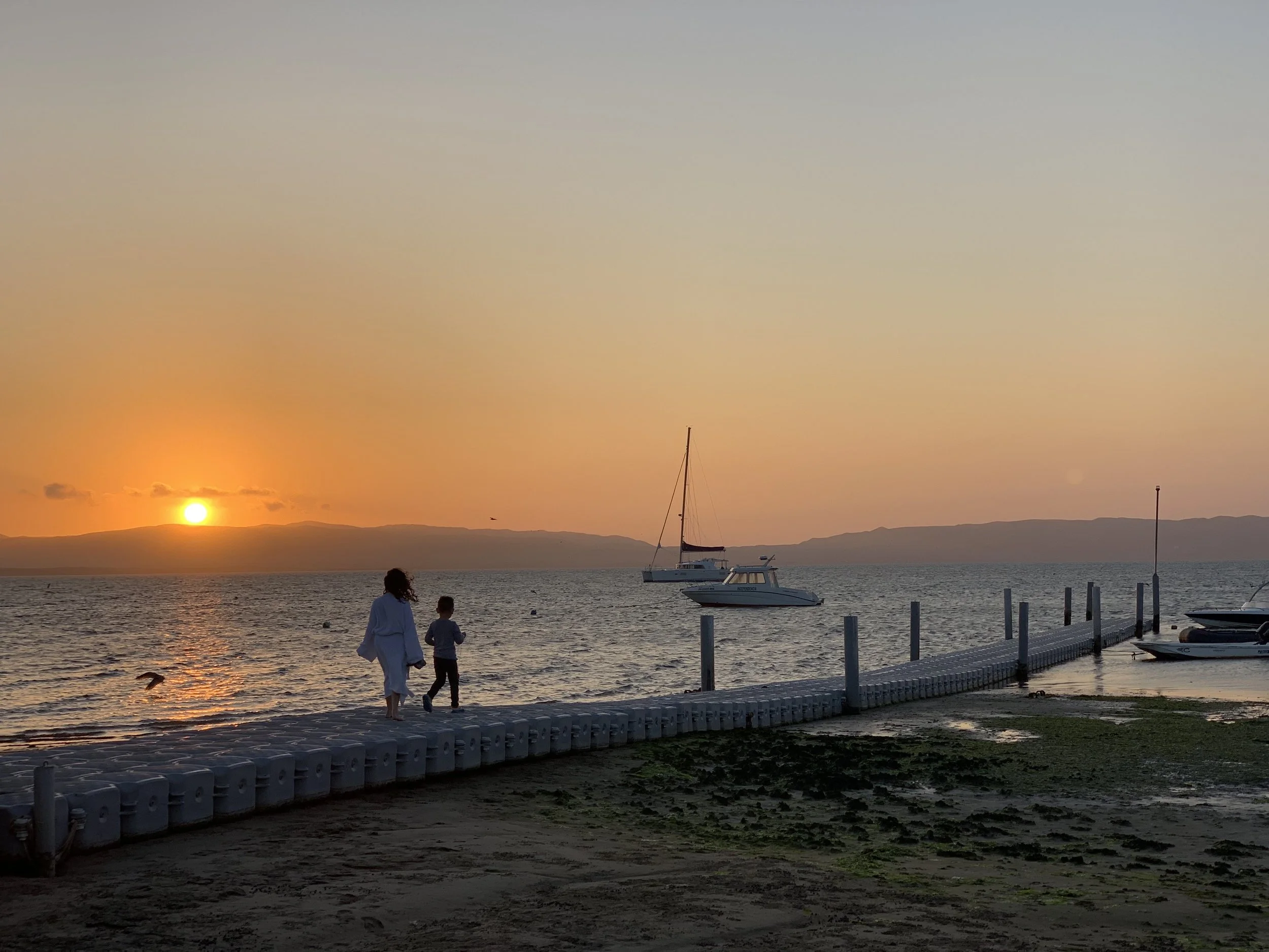 People walking along a shoreline at sunset with sailboats anchored nearby and mountains in the background.
