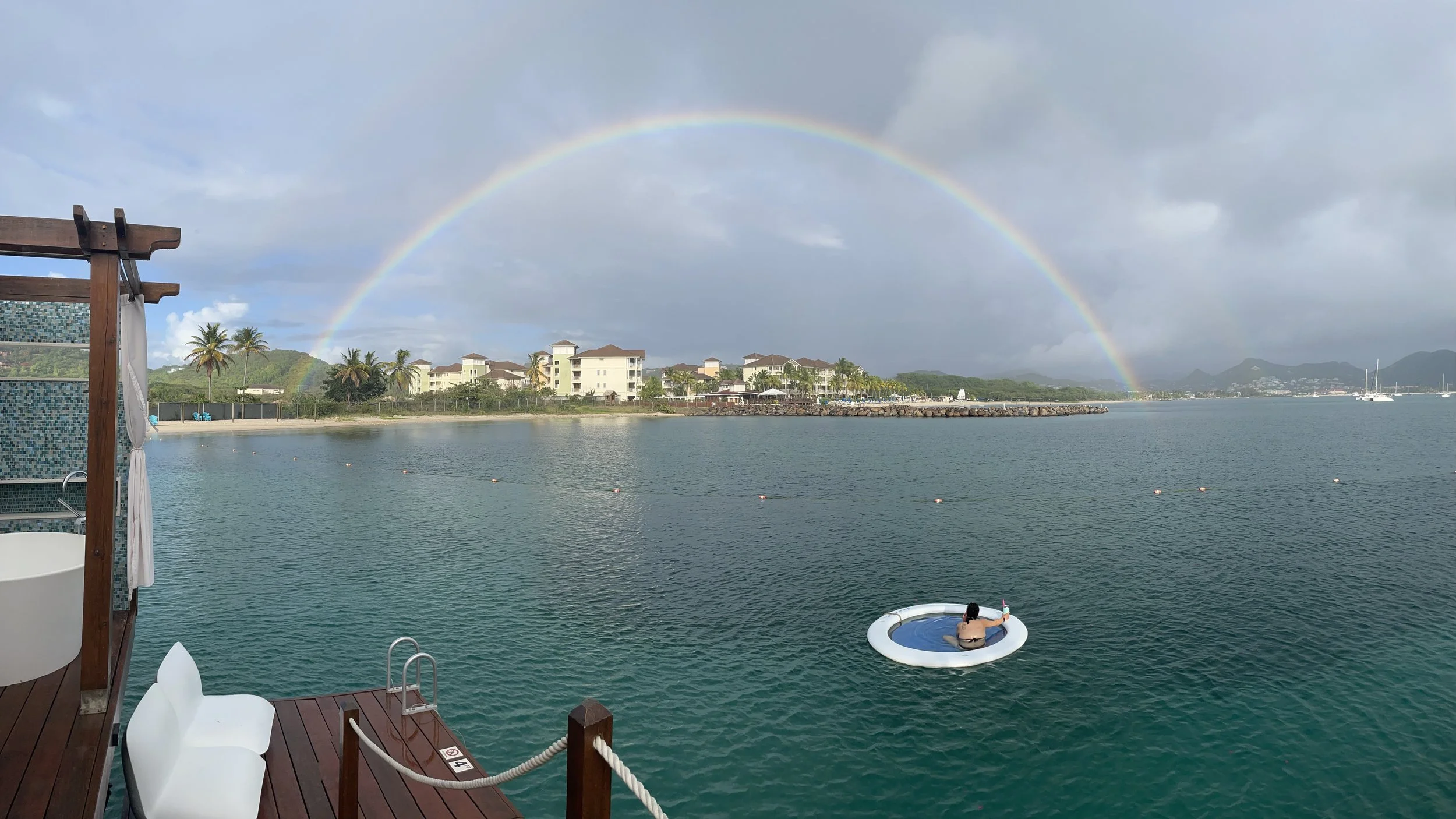 A waterfront scene with a rainbow in the cloudy sky, buildings with palm trees along the shore, and a person relaxing in a circular hot tub on a dock.
