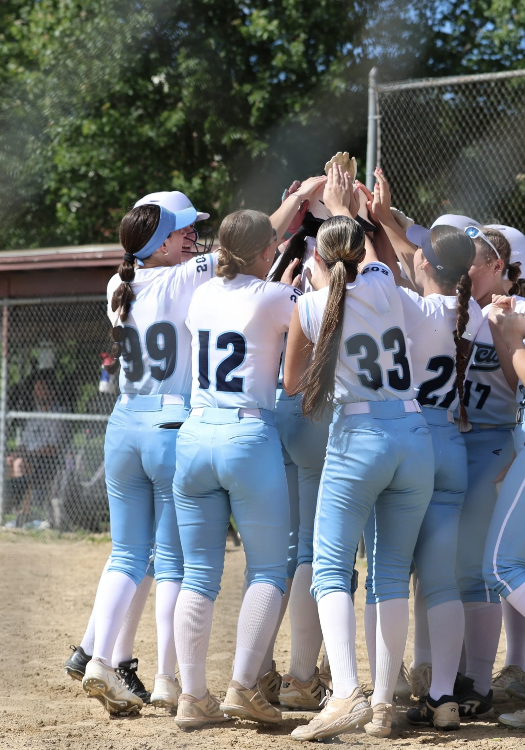 A group of young women softball players in white and blue uniforms celebrating during a game with their hands stacked together.