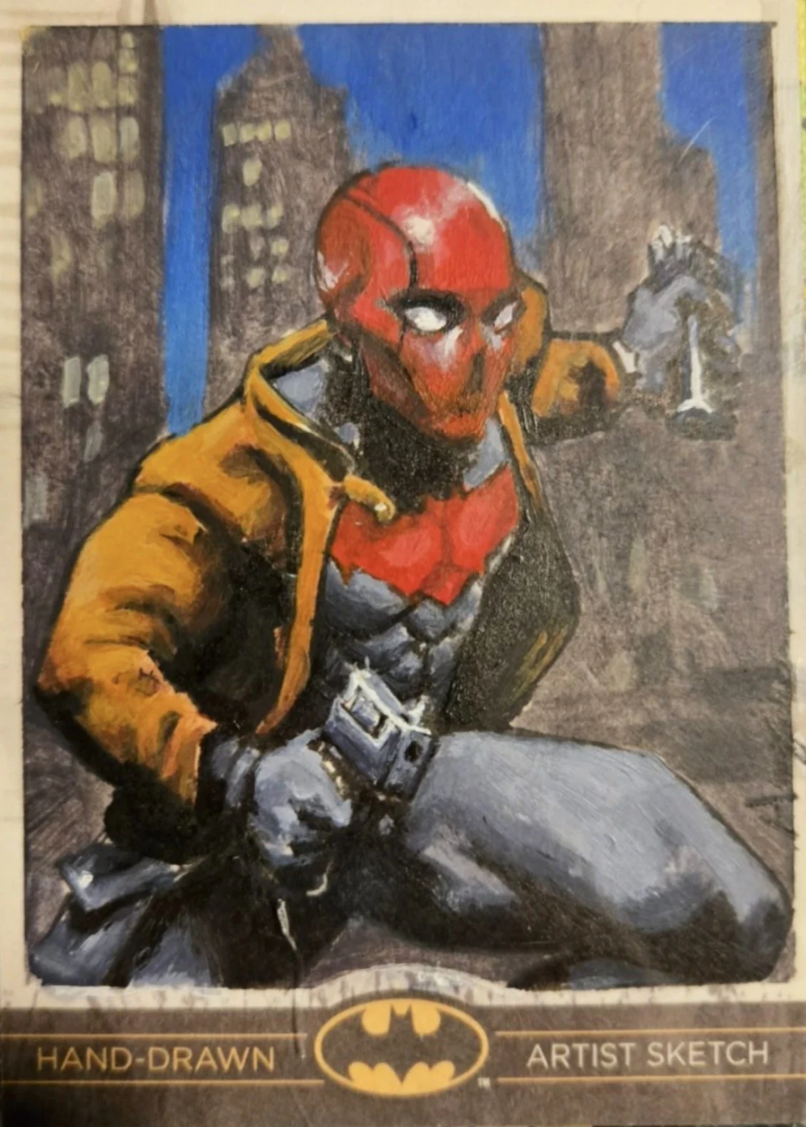 Batman legend sketch card. A hand-drawn artist sketch of Deadpool in a cityscape background, sitting with one knee up, wearing his red and black suit and a yellow jacket, with Batman logo at the bottom.