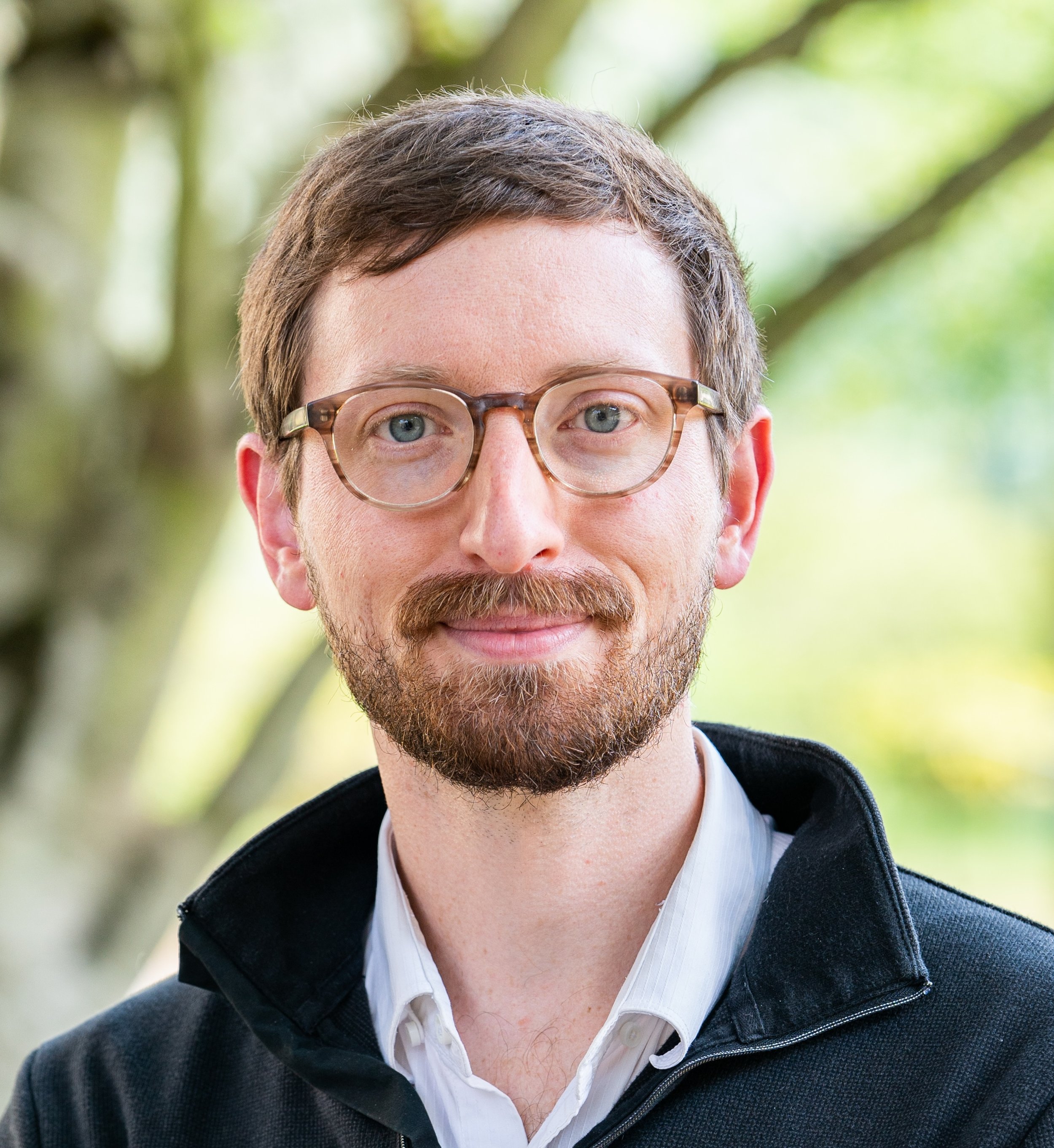 Close-up of a man with light brown hair, a beard, and glasses, wearing a white shirt and black jacket, outdoors with blurred green trees in the background.