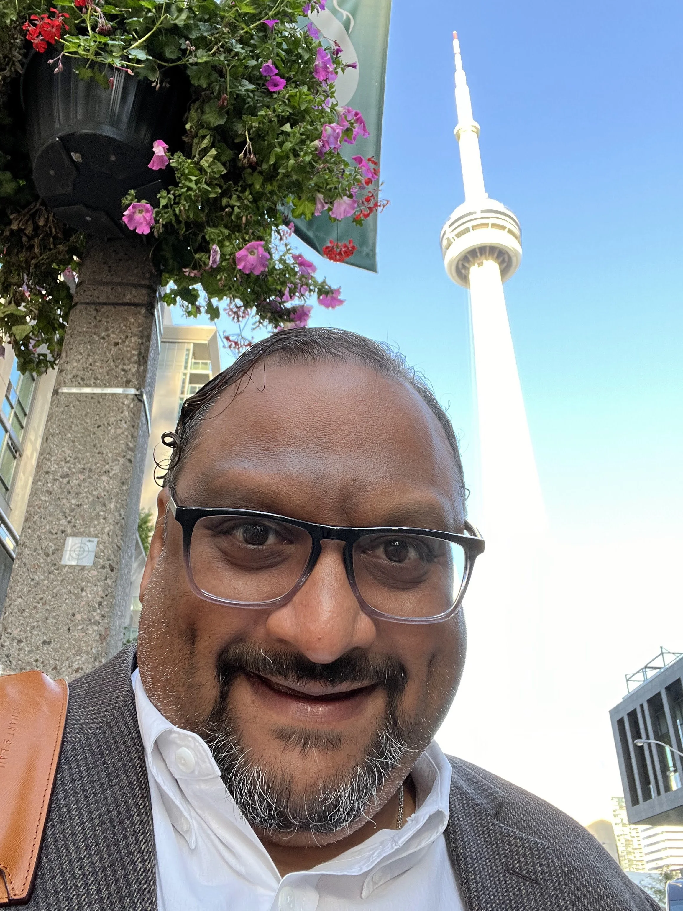 Close-up of a smiling man with glasses and a beard, wearing a white shirt and a brown jacket, taking a selfie outdoors. There are pink and purple flowers in a flower box, a streetlamp, modern buildings, and a clear blue sky in the background.