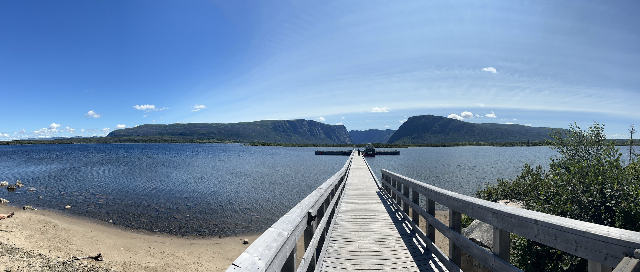 A wooden pier extending into a large body of water with mountains in the distance under a partly cloudy sky.