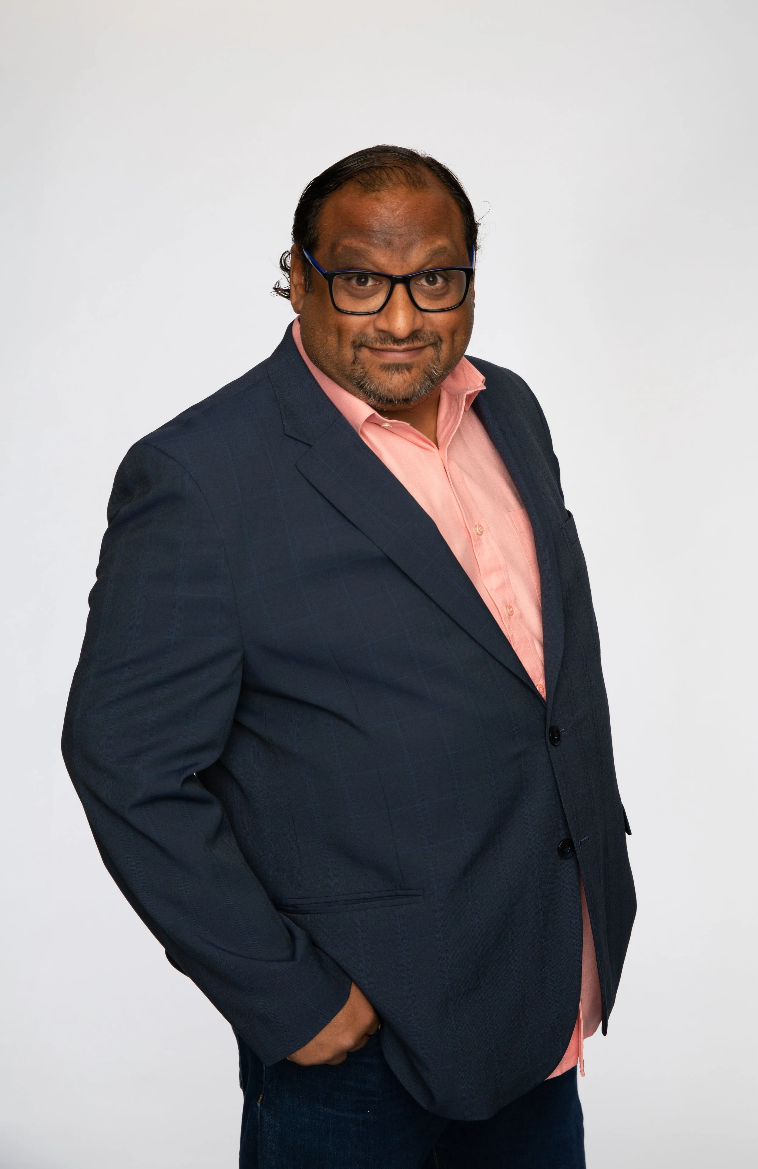 A middle-aged man with glasses, wearing a dark blue suit and pink shirt, stands against a plain white background.